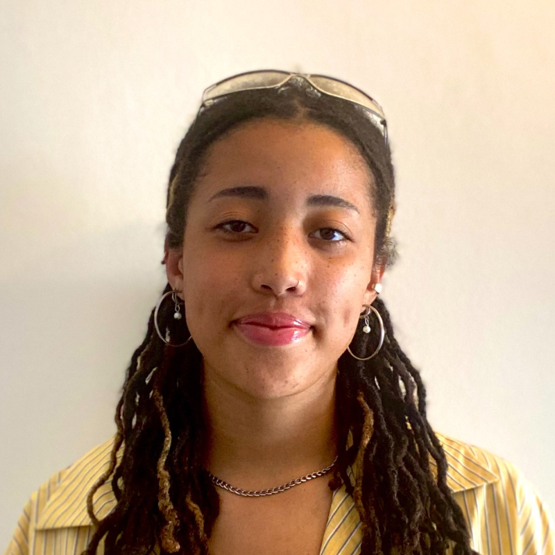 A young woman with dreadlocks wearing gold earrings, a necklace, and glasses on her head, smiling against a plain light-colored background.