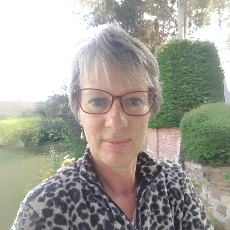 A woman with short gray hair, red glasses, and a leopard print jacket standing outdoors in a garden with trees and shrubs in the background.