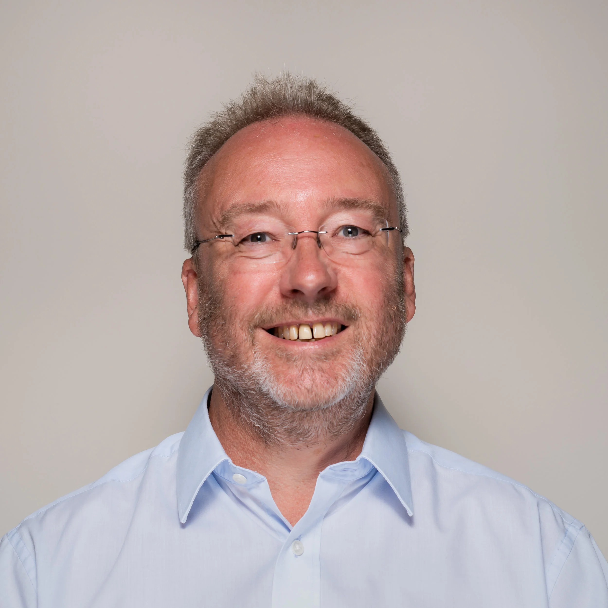 Smiling middle-aged man with glasses and beard, wearing a light blue collared shirt, against a plain neutral background.