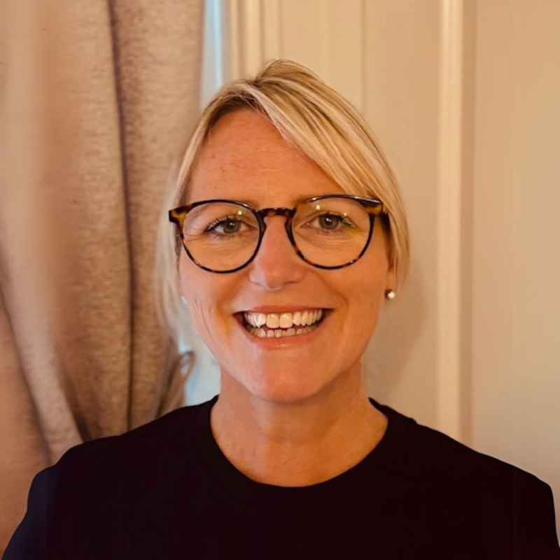A smiling woman with short blonde hair, wearing glasses and earrings, standing indoors with curtains in the background.
