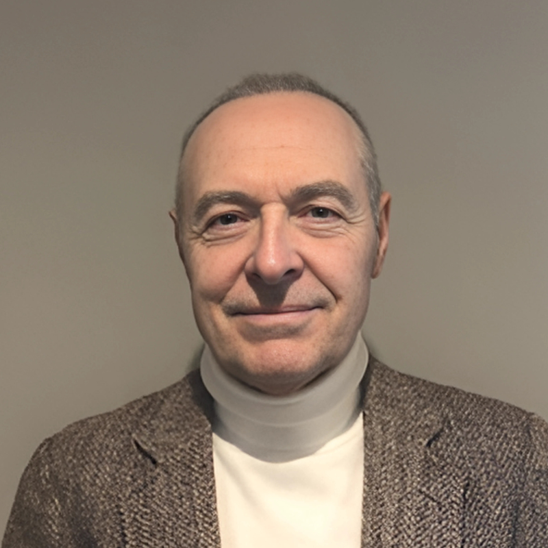A middle-aged man with short gray hair, wearing a tan turtleneck and a brown herringbone blazer, smiling at the camera against a plain gray background.