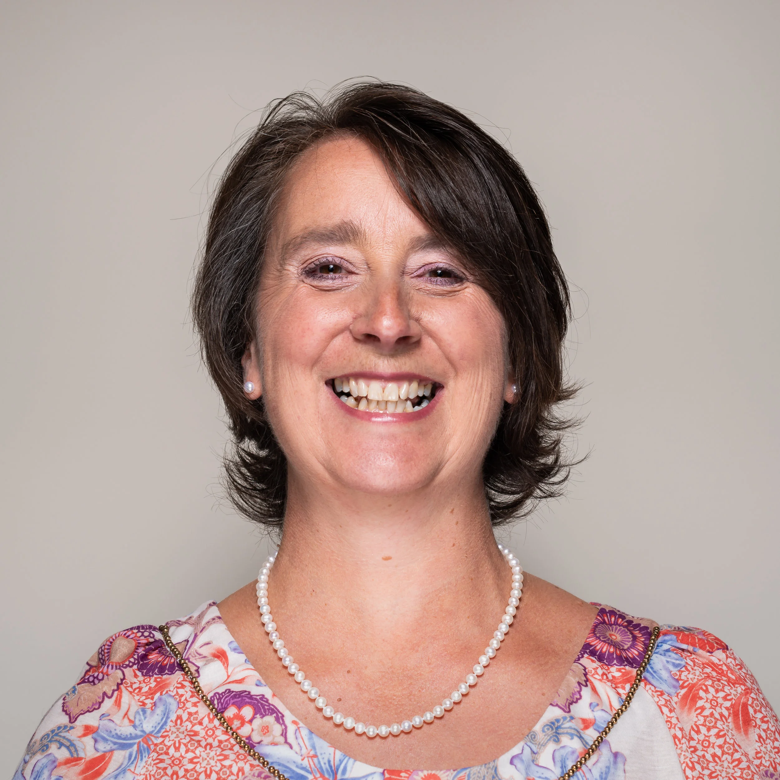 Smiling middle-aged woman with short dark brown hair, wearing pearl earrings, a pearl necklace, and a colorful floral top with orange, purple, and blue patterns, against a plain beige background.