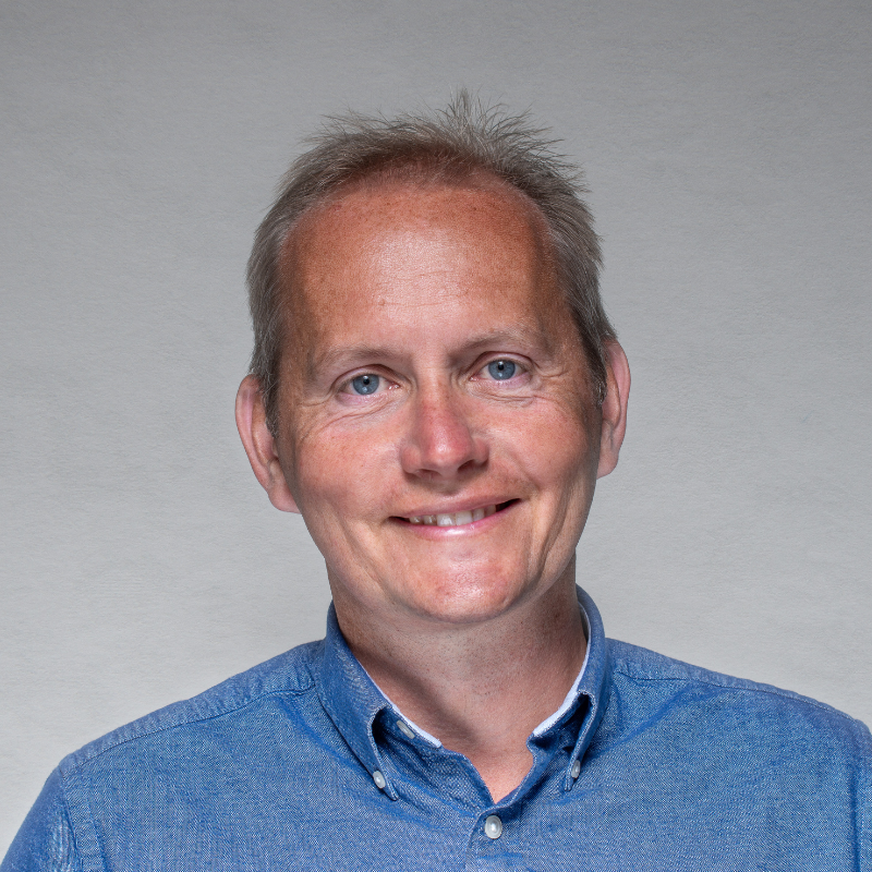 Portrait of a middle-aged man with short light brown hair, blue eyes, and a friendly smile, wearing a blue button-up shirt, against a plain gray background.