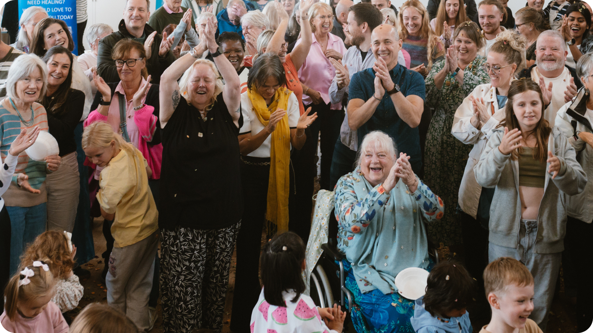A diverse group of people, including seniors, adults, teens, and children, are gathered indoors, smiling, clapping, and celebrating together.