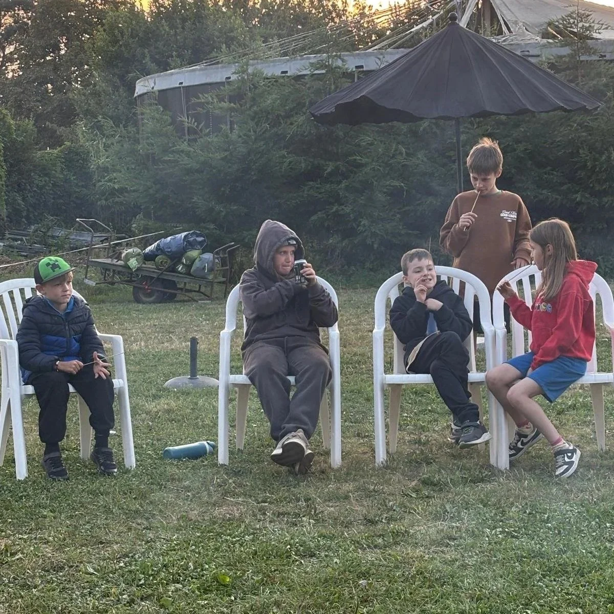 Five children sitting and standing outdoors near white chairs, some holding sticks or drinks, with an umbrella and trees in the background.