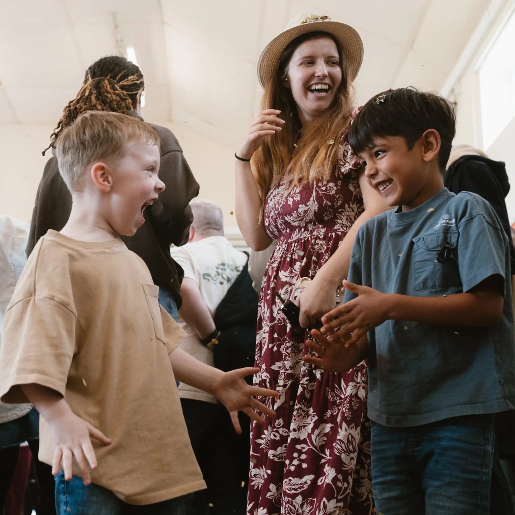 A group of children and a woman laughing and talking indoors, with others blurred in the background.