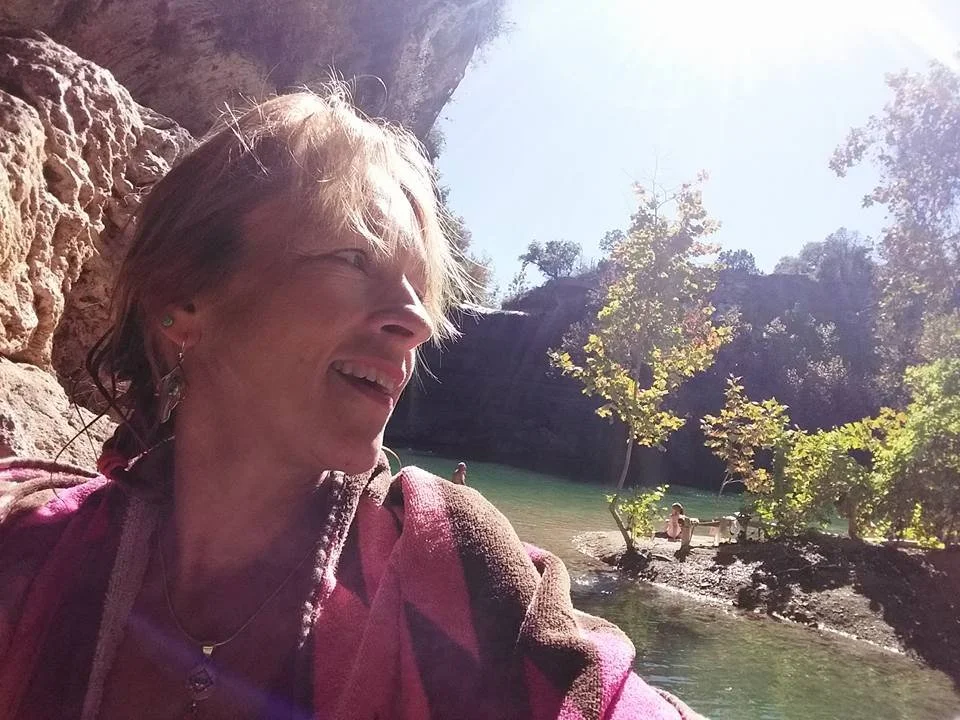 A woman with short light hair smiling and looking to the side, sitting outdoors near a lush river with trees and rocks, sunlight shining through the trees.