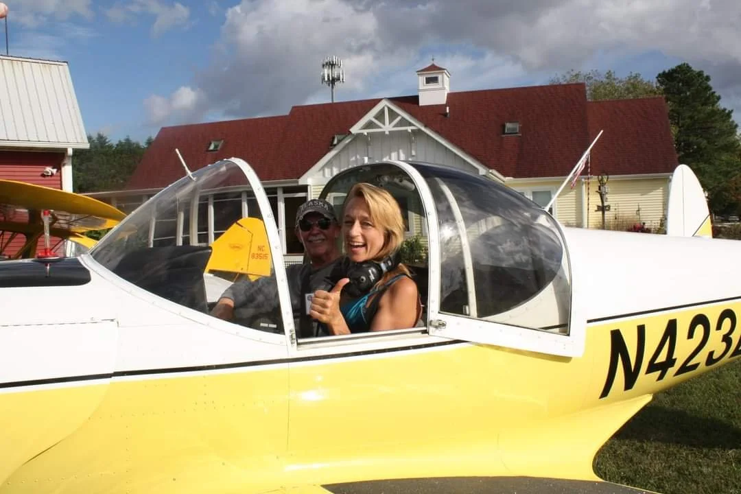 Two people sitting inside the cockpit of a yellow aircraft, smiling and giving a thumbs-up, with a house and blue sky with clouds in the background.