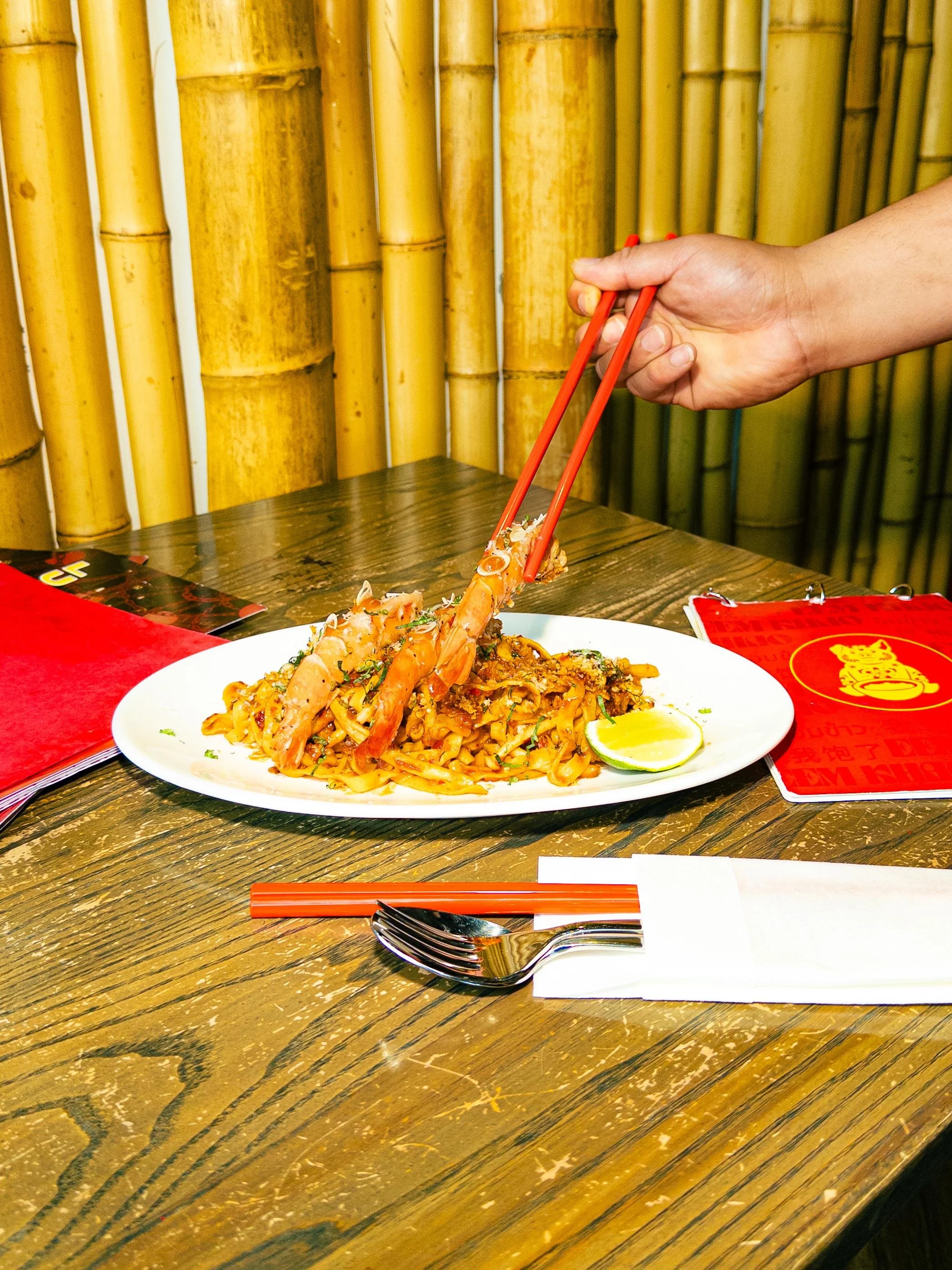 Hand using red chopsticks to pick up a shrimp from a plate of Asian noodles garnished with vegetables and lime wedge on a wooden table.