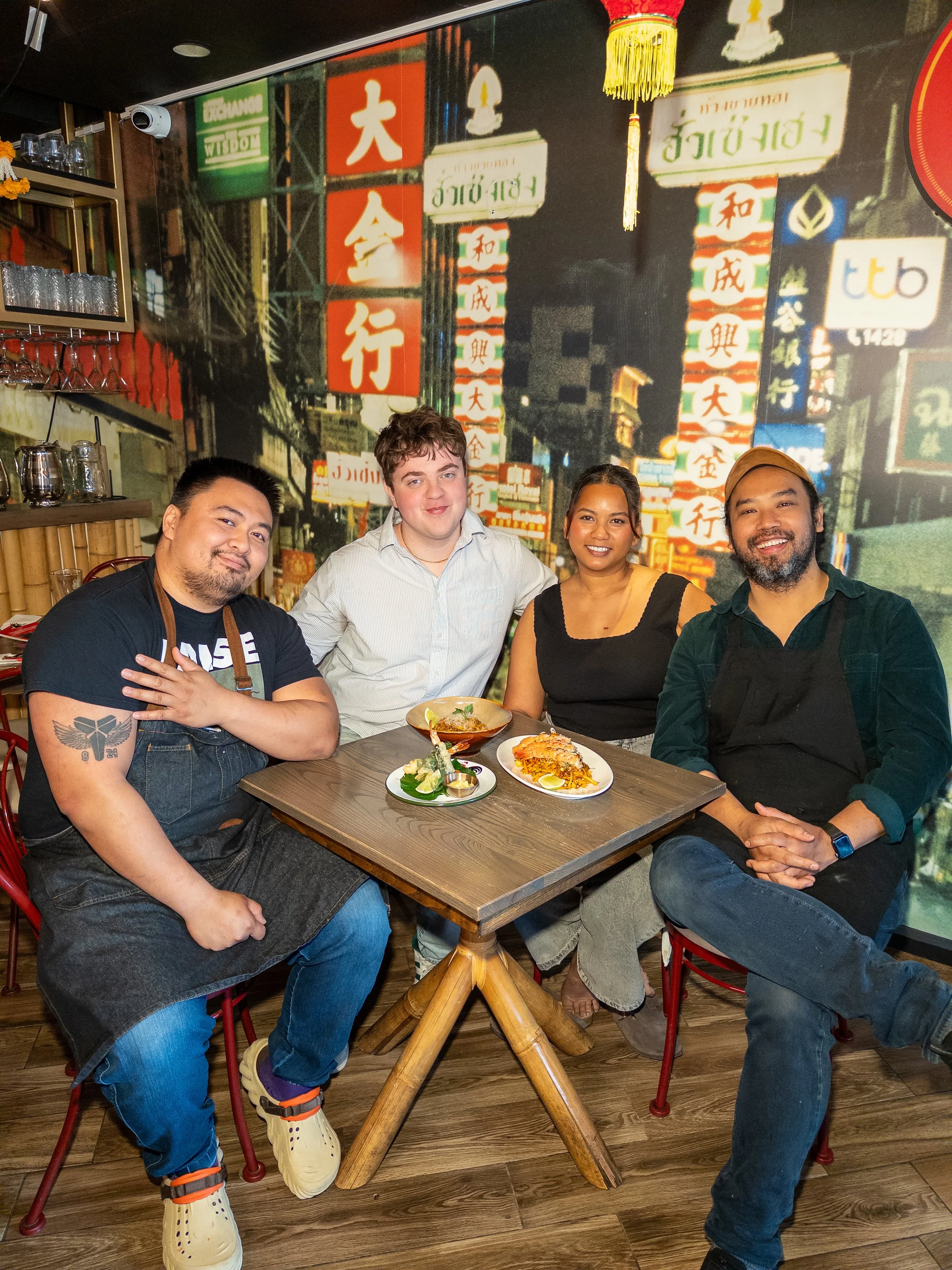 Four people sitting around a dining table in a restaurant with Asian decor, dishes of food on the table, and a colorful cityscape mural in the background.