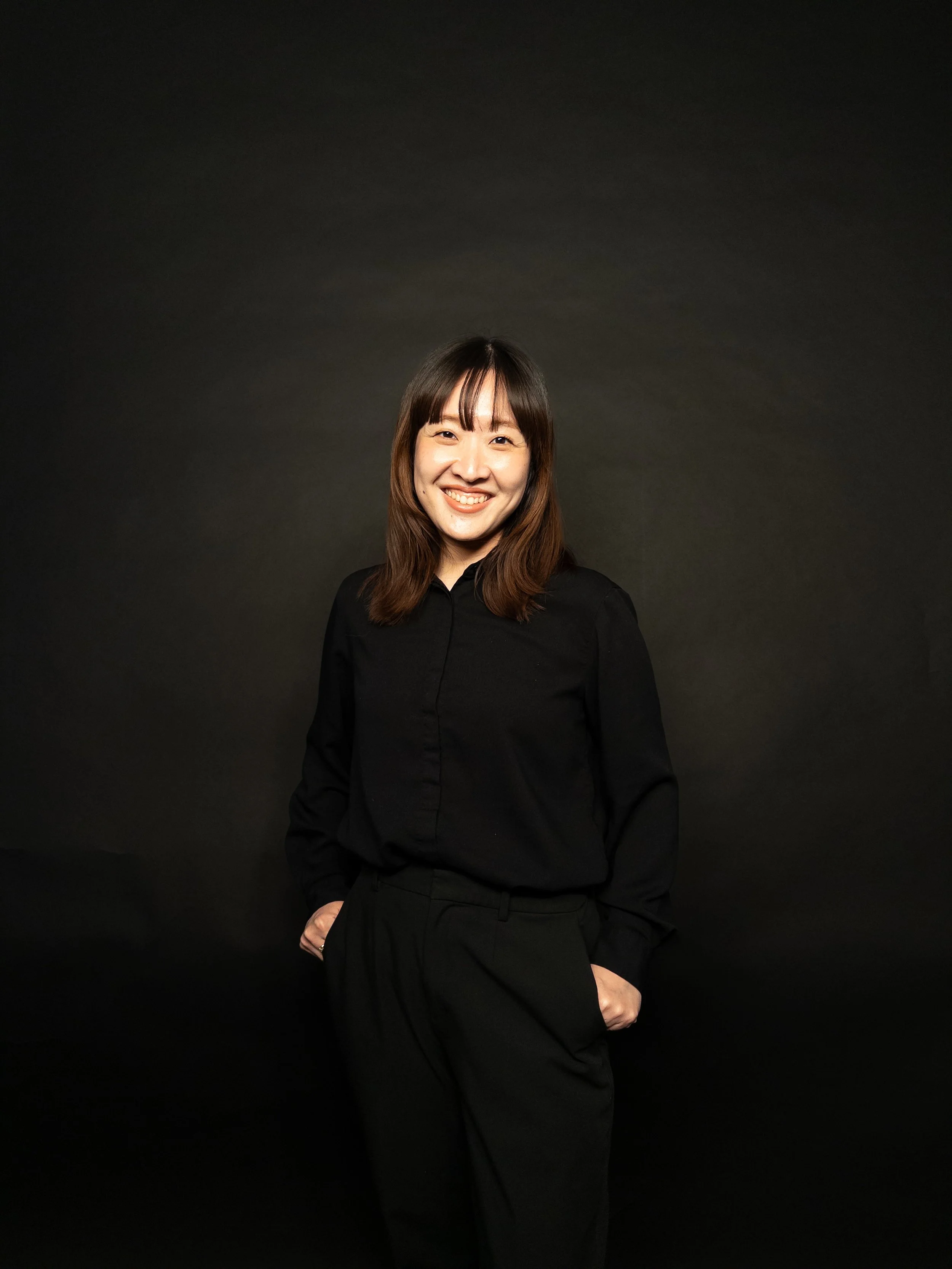 A young woman with shoulder-length brown hair, wearing a black shirt and black pants, smiling and standing against a plain dark background.