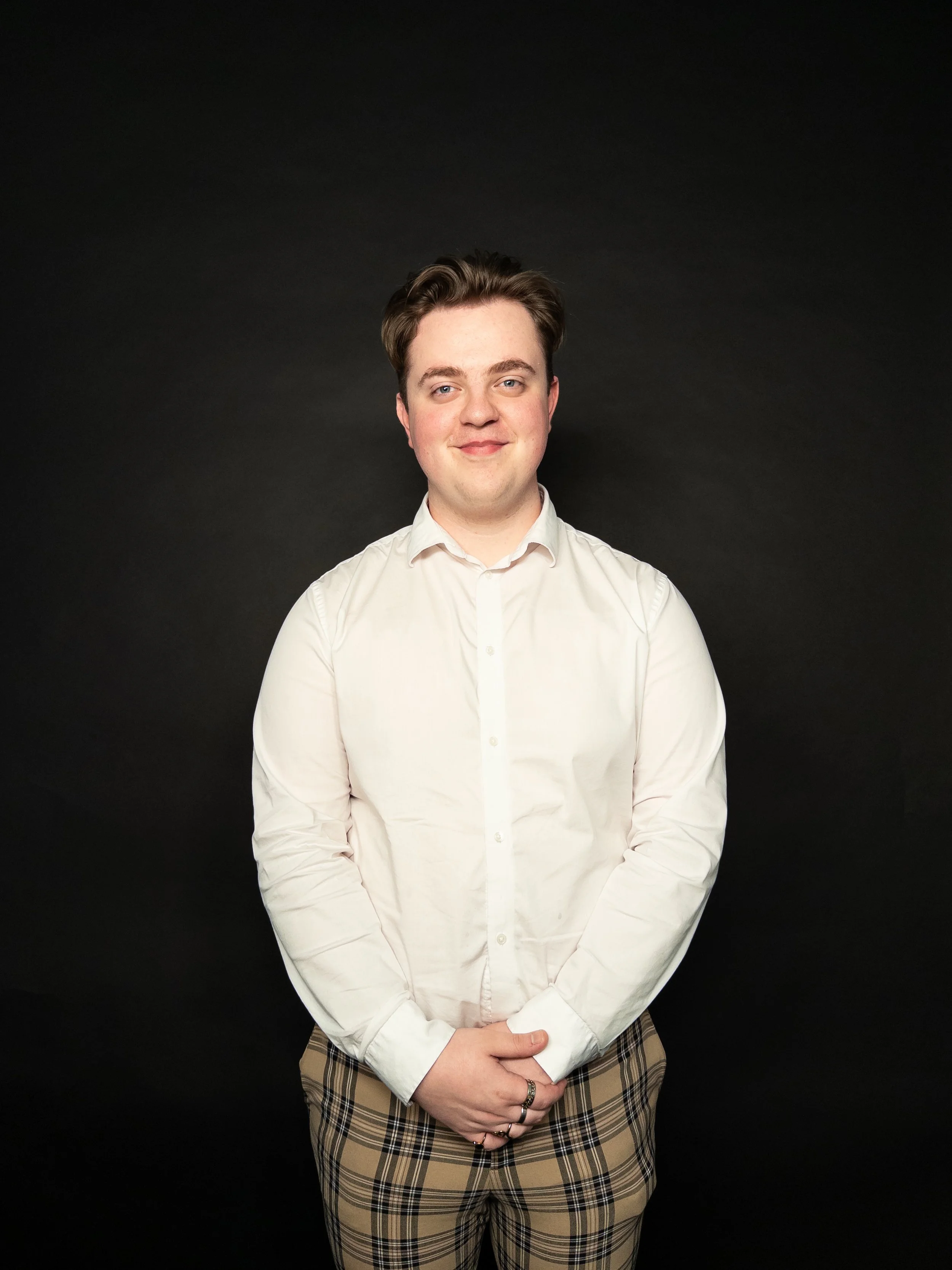 A young man with brown hair, wearing a white button-up shirt and beige plaid pants, standing against a dark background and smiling.