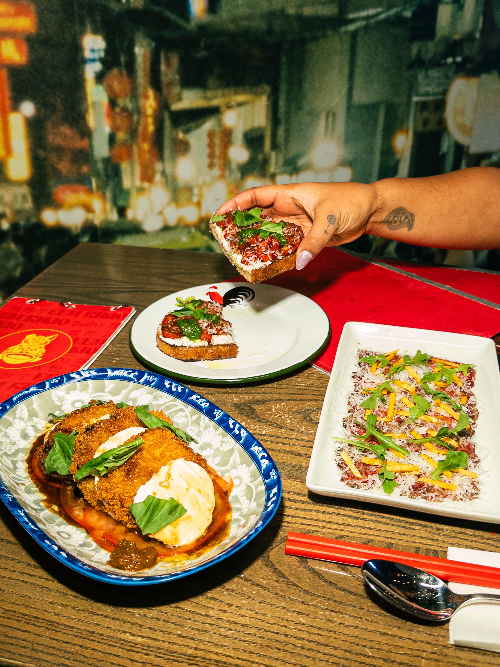 Three dishes including a pizza slice topping with greens, a breaded dish with a poached egg, and a plate with thinly sliced meat garnished with greens and shredded cheese; a person's hand holding the pizza slice with a cityscape background.