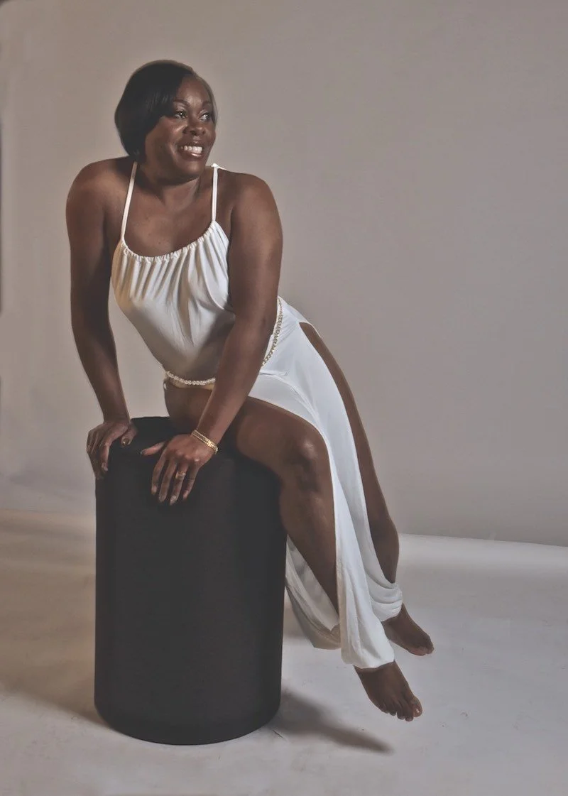 A woman in a white dress sitting on a black cylindrical stool, smiling and looking to her left against a plain background.