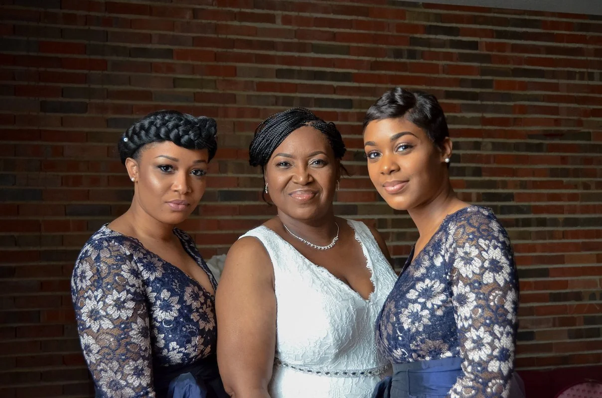 Three women in lace dresses posing against a brick wall, smiling softly.