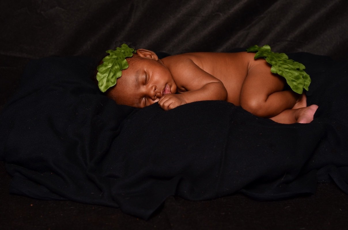A young child is sleeping peacefully, lying on their side on a black fabric surface with a black cloth underneath them. The child has two large green leaves placed on their head and on their back.