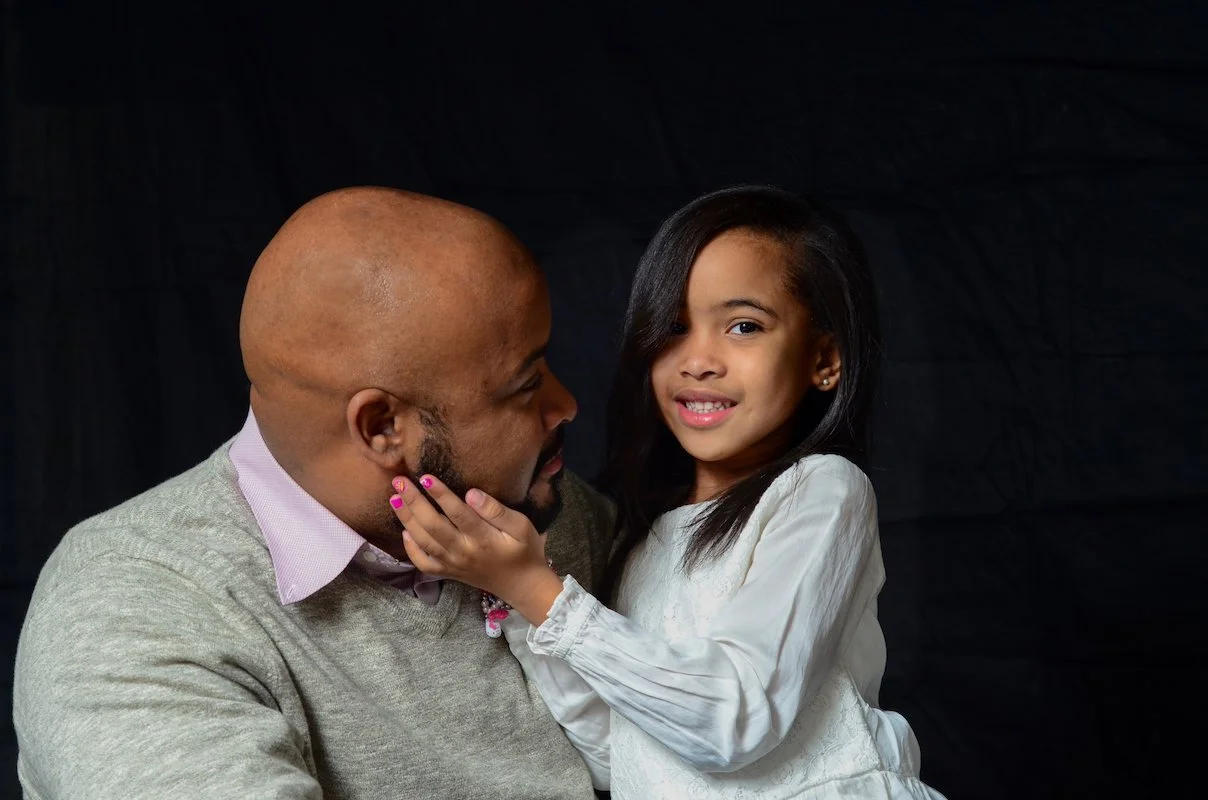 A man and a young girl share a tender moment against a black background. The girl is gently cupping the man's face with her hand, and they are looking at each other with affection.