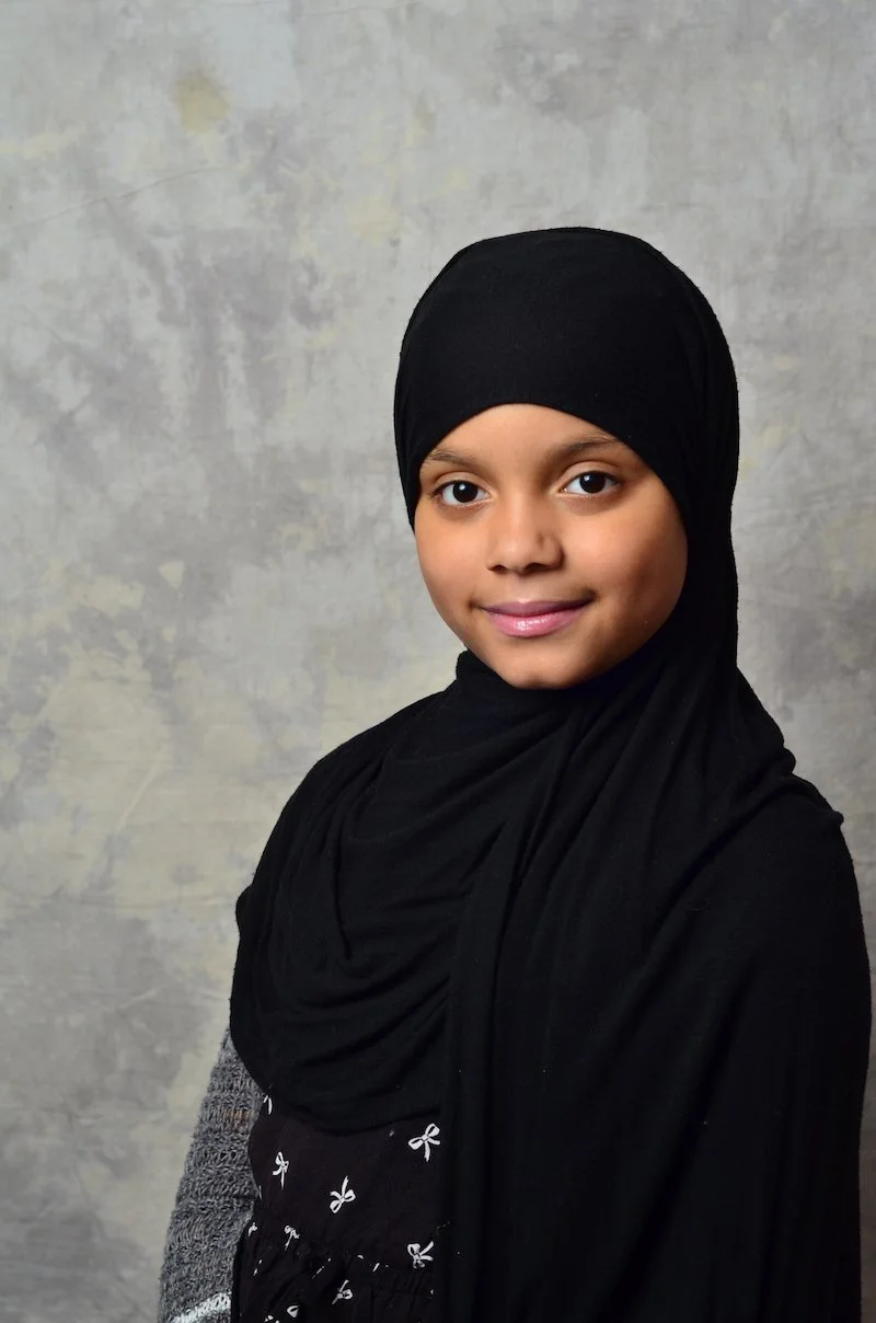 A young girl wearing a black hijab, smiling softly, against a gray textured background.