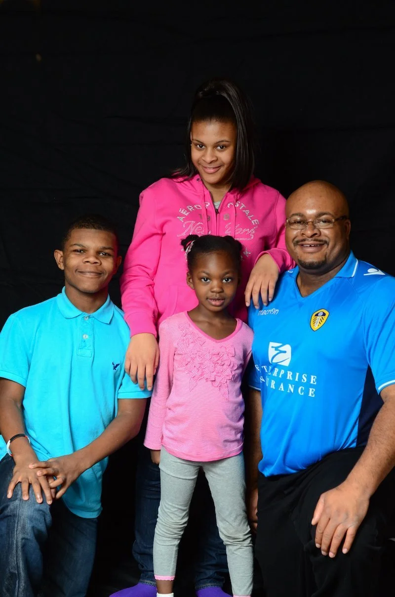 A group of four African American children and one adult man smiling and posing together against a black background. The girl at the top is wearing a pink hoodie, the girl in the middle is in a pink shirt, and the boy at the left is in a light blue polo. The man on the right is wearing a blue sports jersey with a yellow emblem and sponsor logos.
