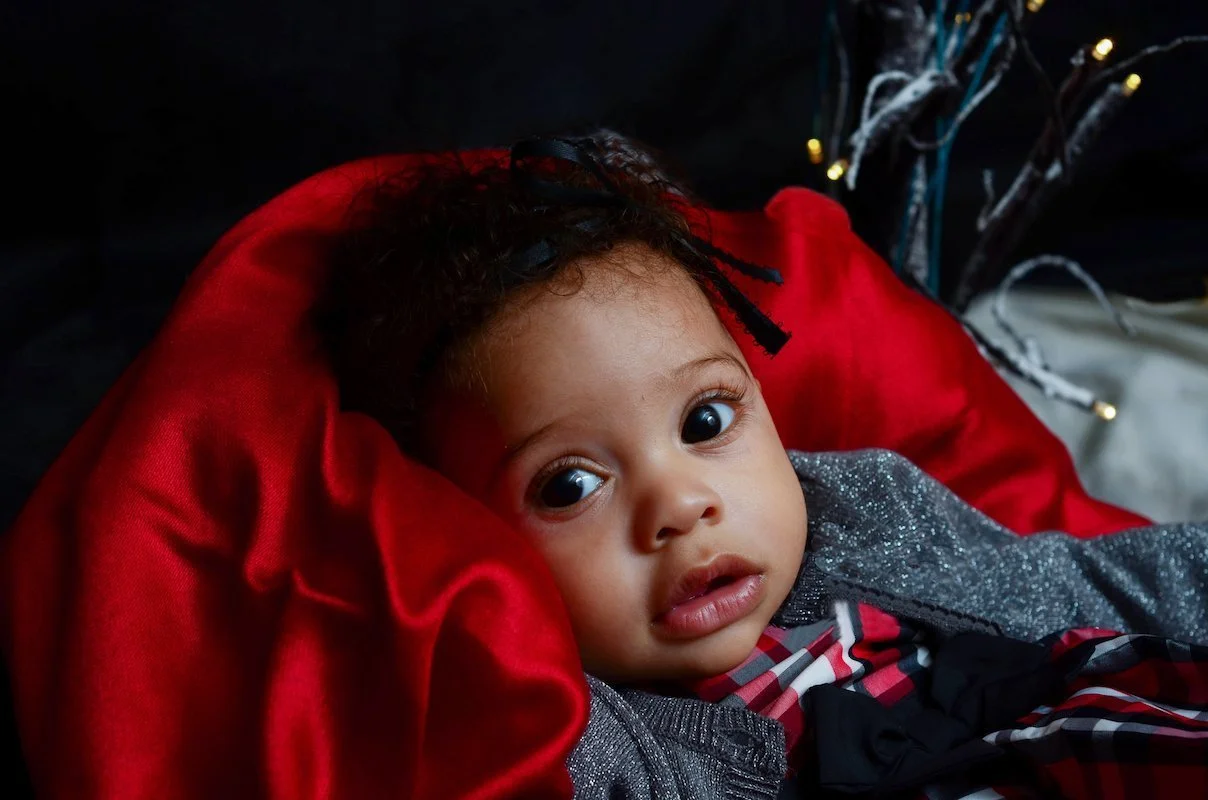 A young child with curly hair and big eyes lying on a plush red pillow, wearing a gray, red, and black plaid outfit, with faint holiday lights in the background.