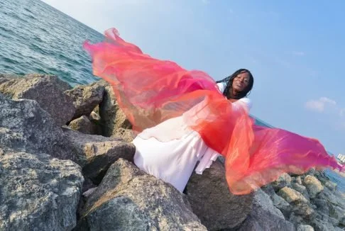 Woman in a white dress holding a colorful sheer fabric on a rocky shoreline with the ocean and blue sky in the background.