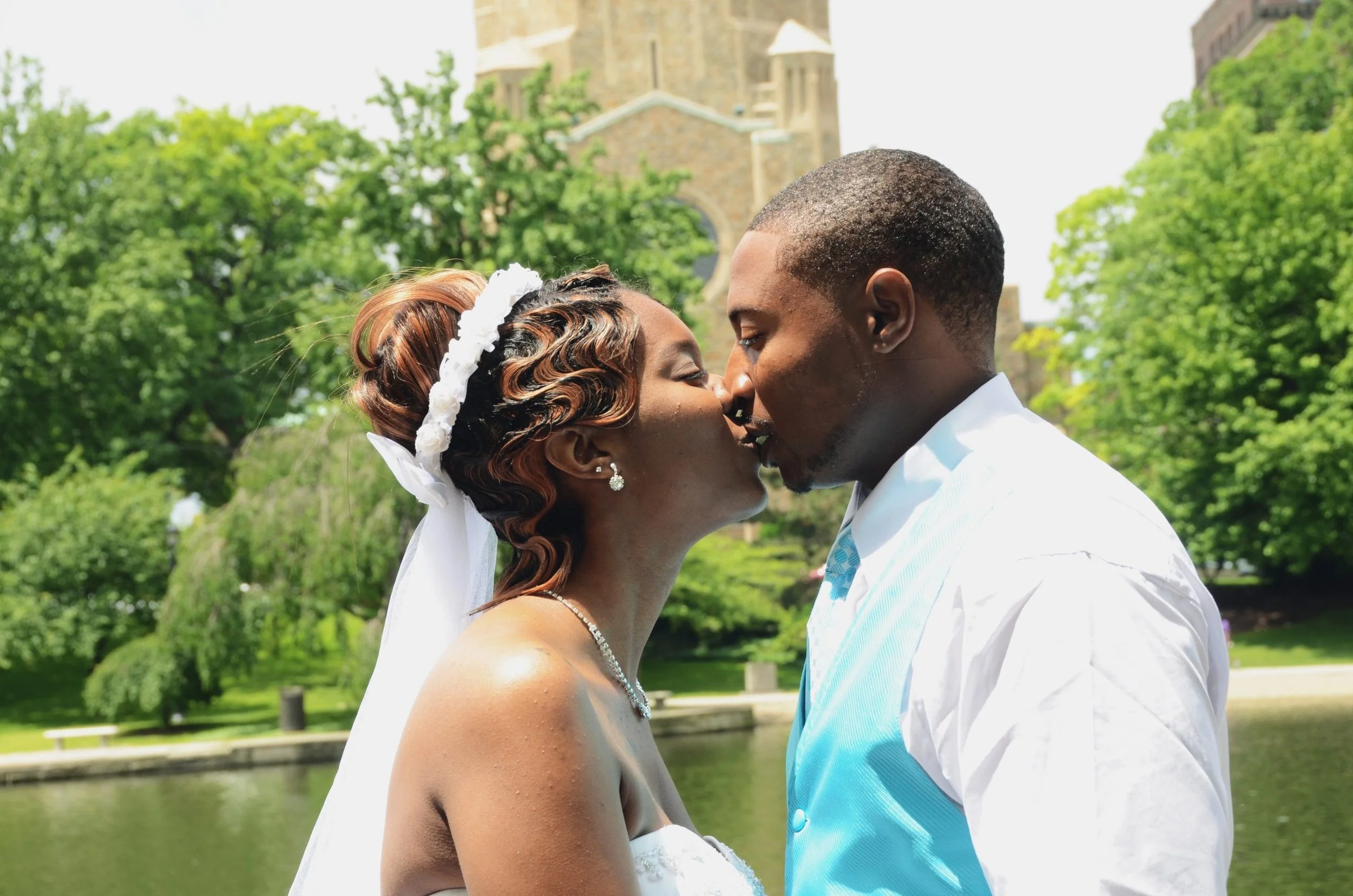 A bride and groom sharing a kiss outdoors during their wedding, with green trees and a historic building in the background.