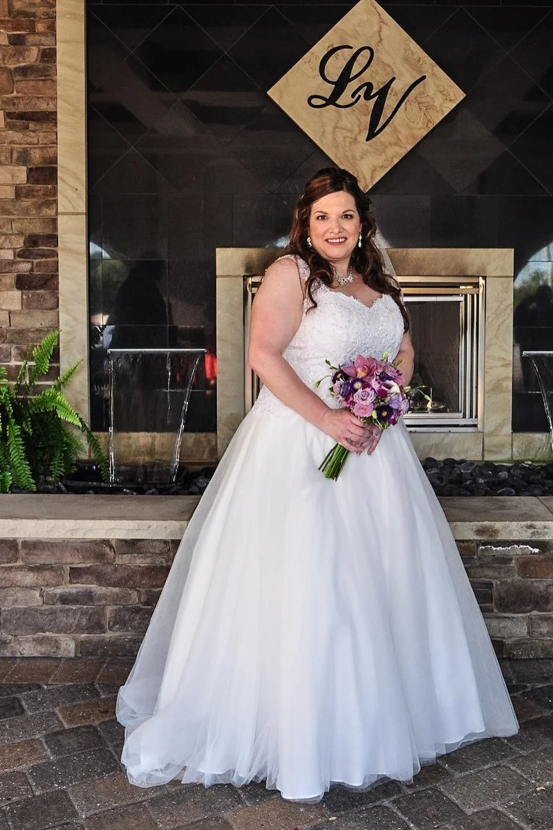 A woman in a white wedding gown holding a bouquet of purple and pink flowers, standing in front of a modern fireplace with black and stone accents, and a decorative sign with the initials 'LyV' above.