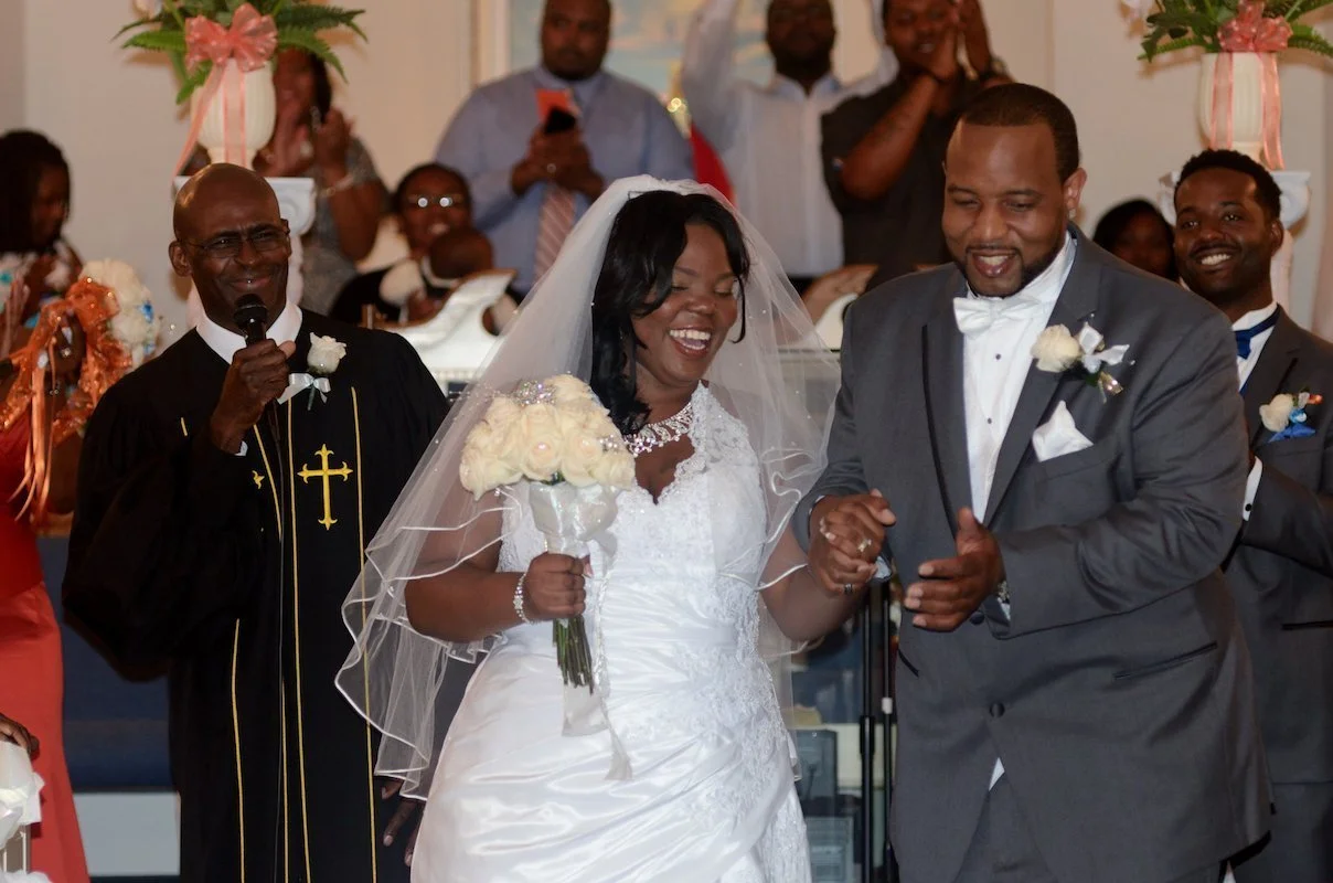 A bride and groom smiling and holding hands during their wedding ceremony in a church, with a priest and guests in the background.