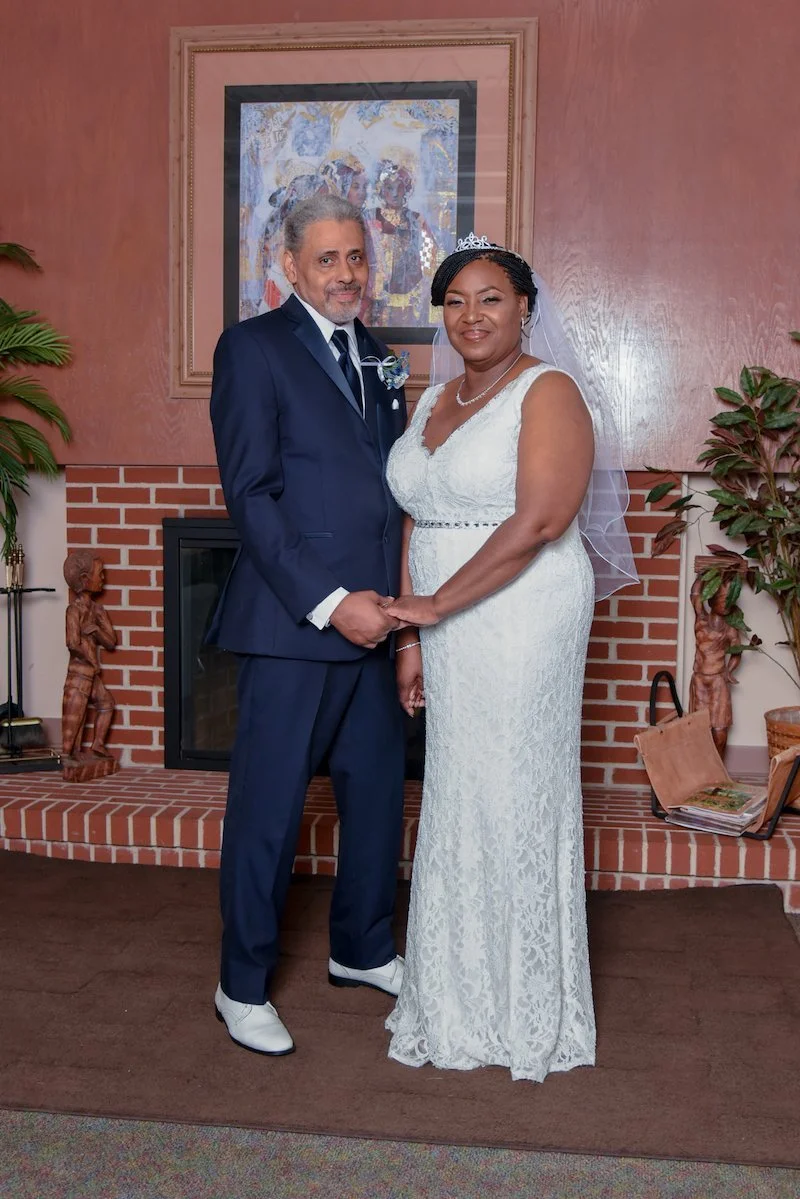 A bride and groom standing together indoors, holding hands, in front of a fireplace with a framed artwork above it. The groom is wearing a blue suit with a tie, and the bride is wearing a white lace wedding dress with a veil and tiara. Both are smili