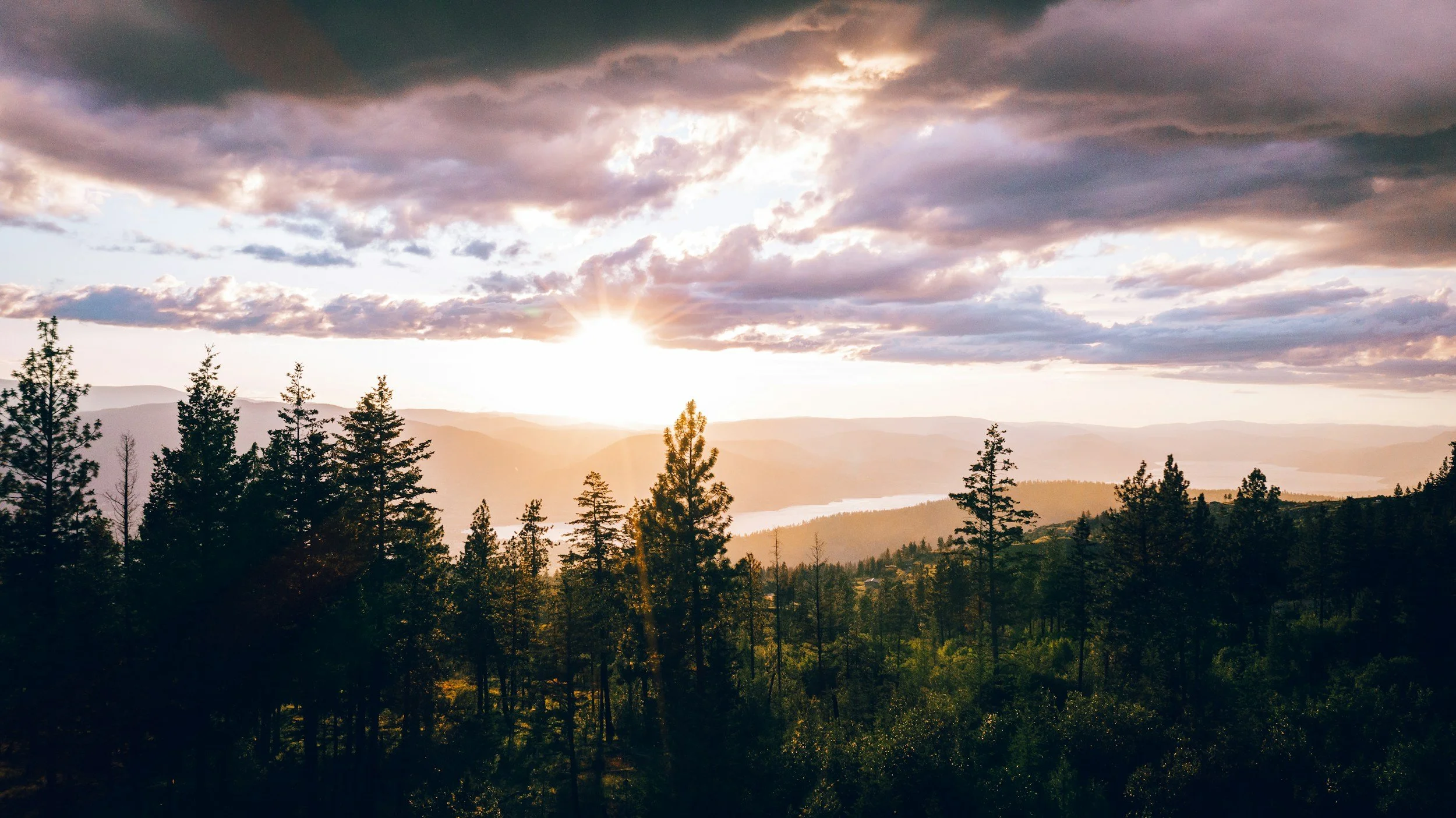Sunset over a forested landscape with rolling hills and a lake in the distance, clouds in the sky