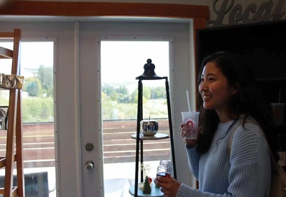 A woman with dark hair smiling and holding a drink with a straw inside a cozy indoor space with large windows, a wooden ladder, and decorative items on shelves.