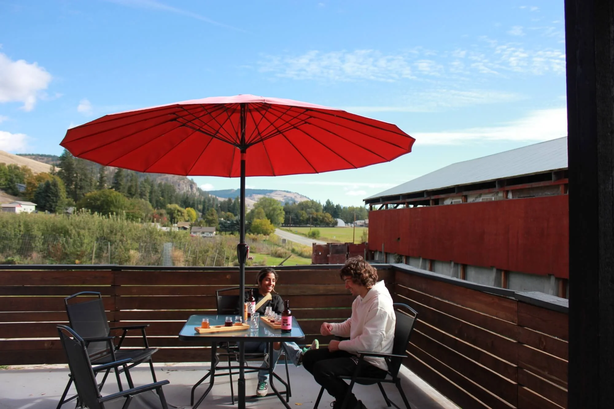 Two people sitting at a table on a patio with a large red umbrella, enjoying a drink outdoors with a scenic view of green hills and blue sky.