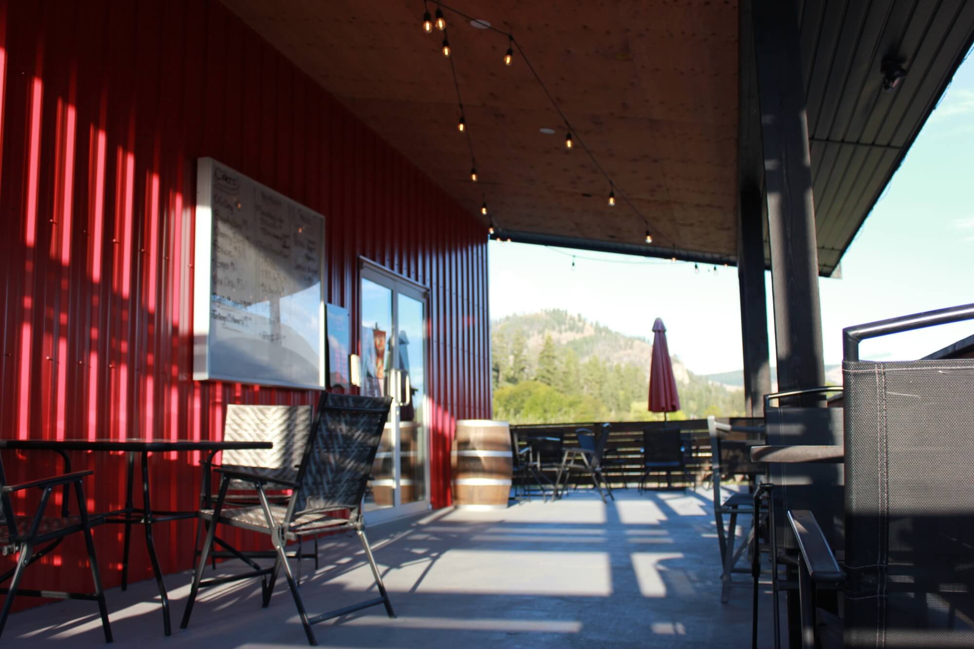 Outdoor patio of a Tony's Craft Cidery with black metal chairs and tables, string lights hanging from the ceiling, a red metal wall with a menu board, and a scenic mountain view in the background.