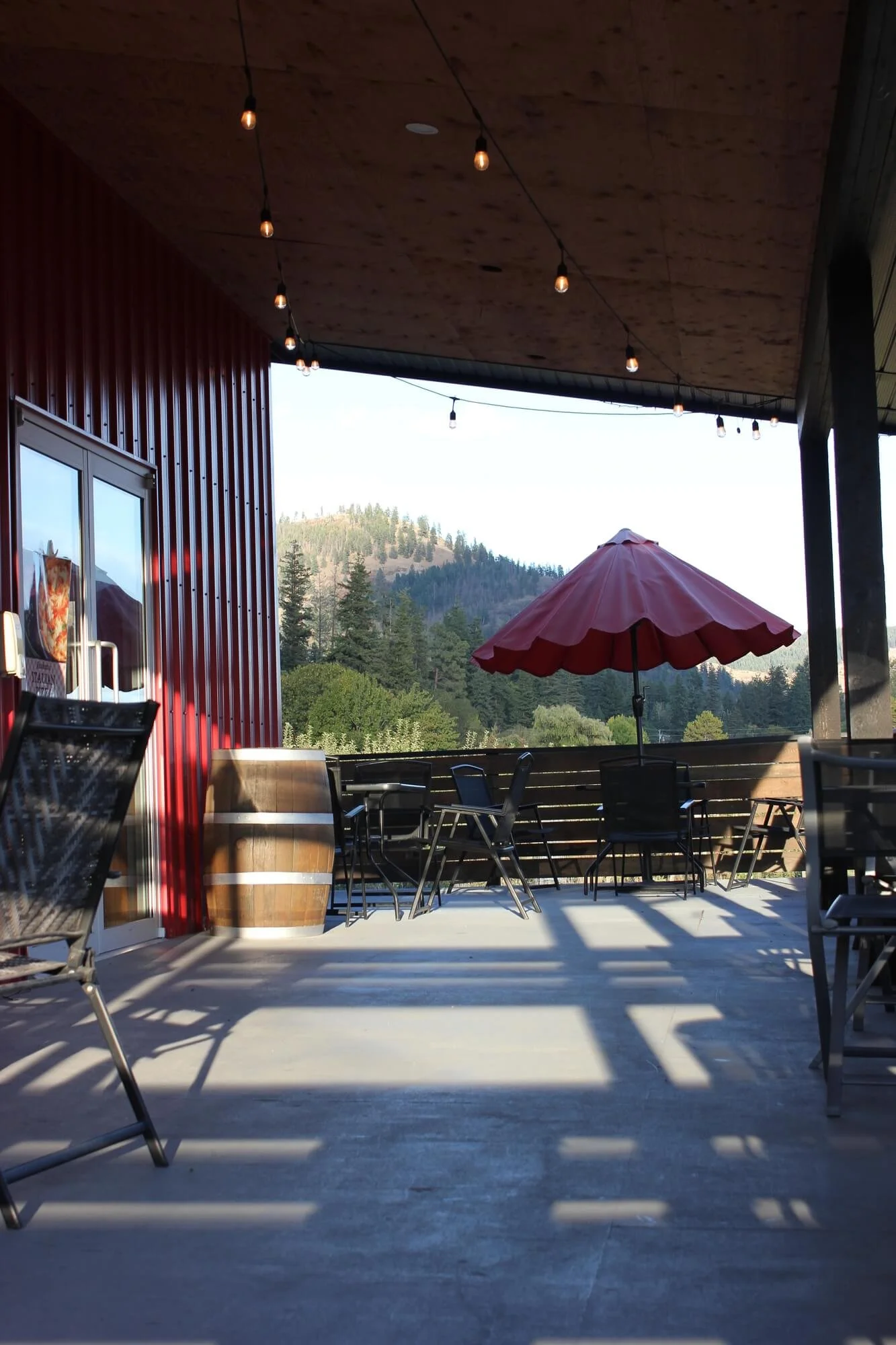 Empty outdoor patio with chairs, a table with an umbrella, a barrel, and string lights, overlooking a mountainous landscape with trees.