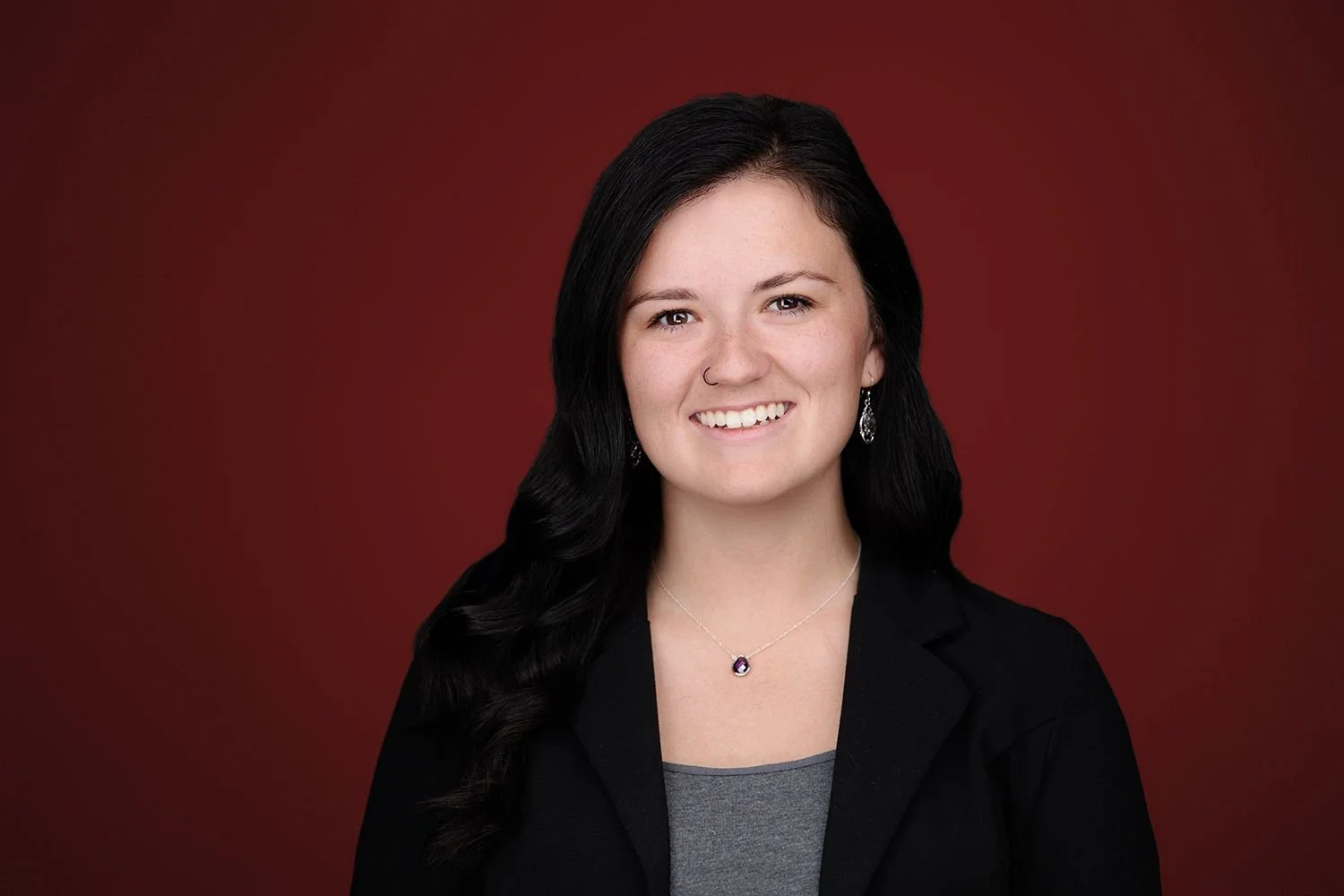 Portrait of a young woman with long dark hair, wearing a black blazer and a silver necklace with a purple gemstone, smiling against a red gradient background.