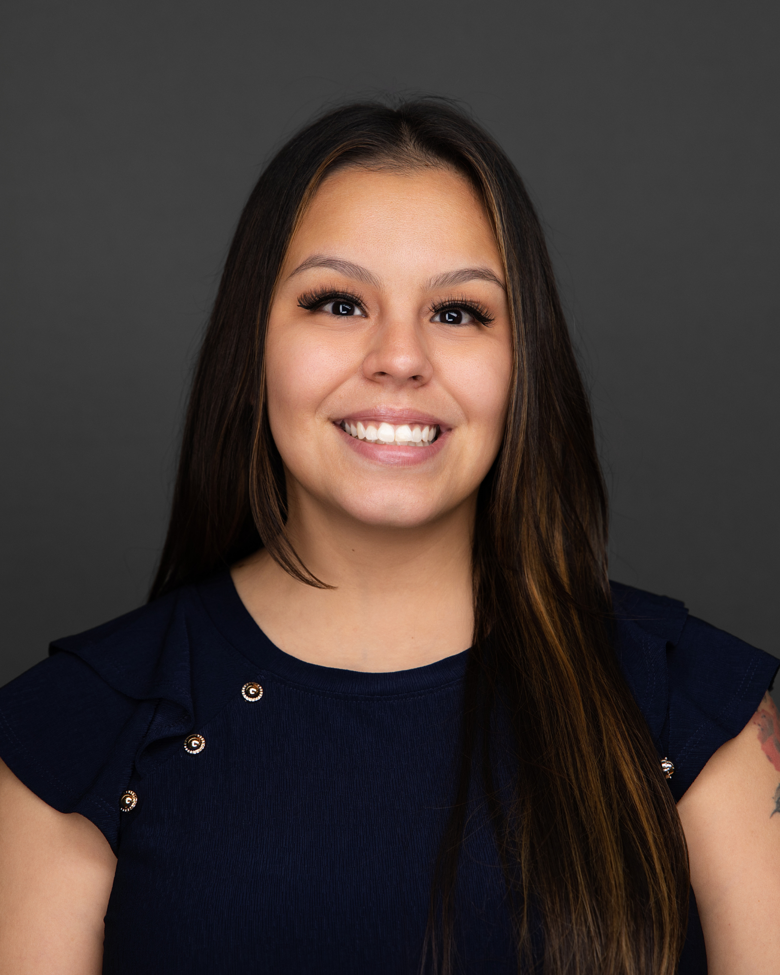 A young woman with long dark hair, wearing a dark navy top with shoulder details, smiling at the camera against a gray background.