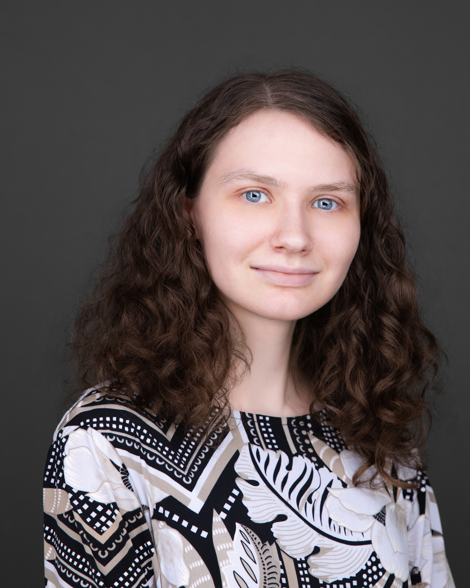 A young woman with long, curly brown hair and blue eyes, wearing a black, white, and beige patterned blouse, against a dark gray background.