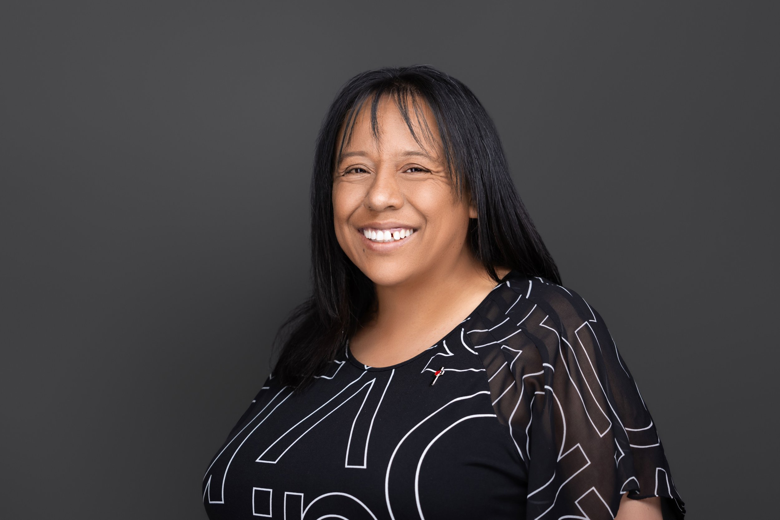 A woman with long black hair smiling, wearing a black shirt with white line art design, against a dark gray background.