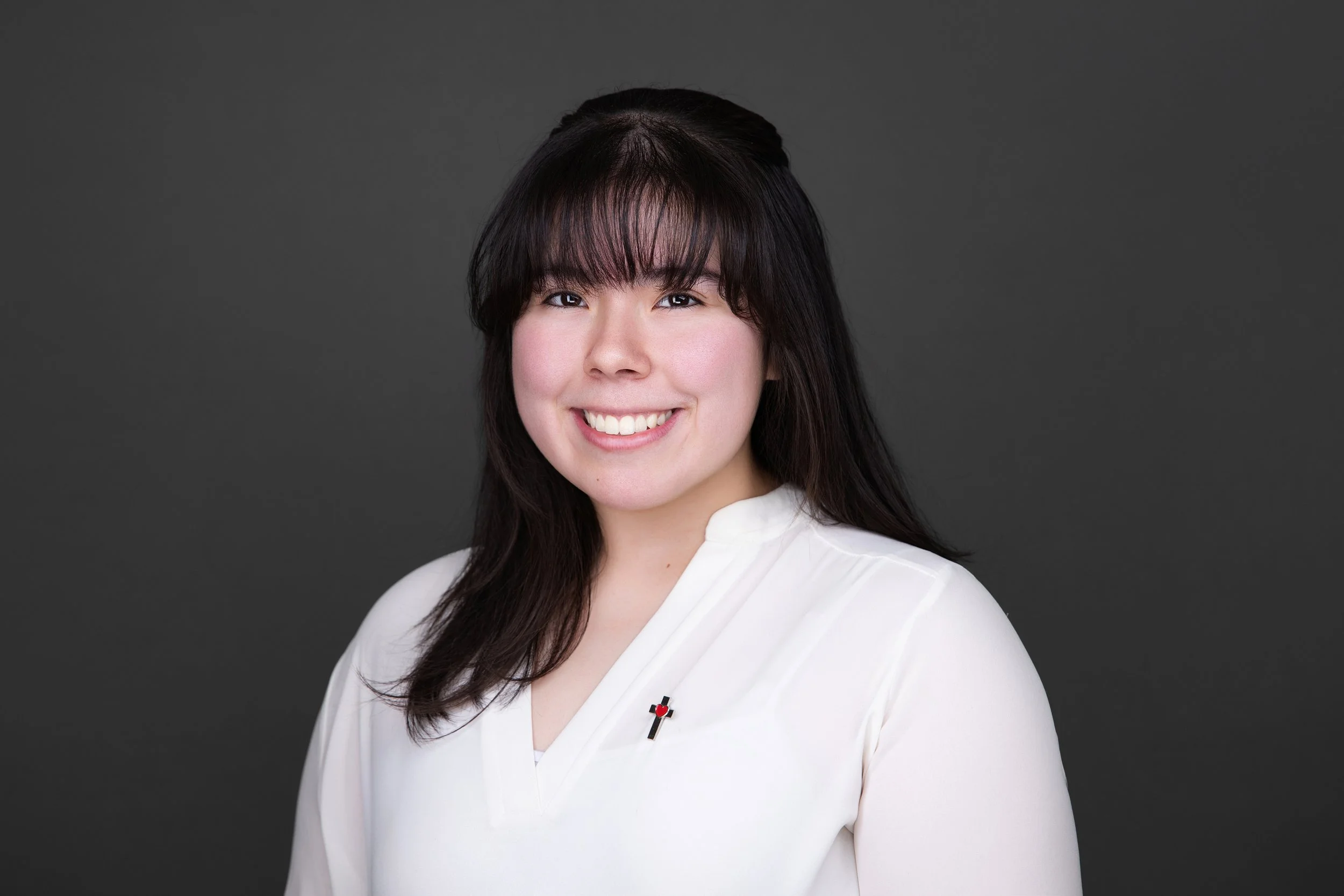 Portrait of a woman with dark hair, wearing a white top with a small cross pin, smiling against a dark gray background.
