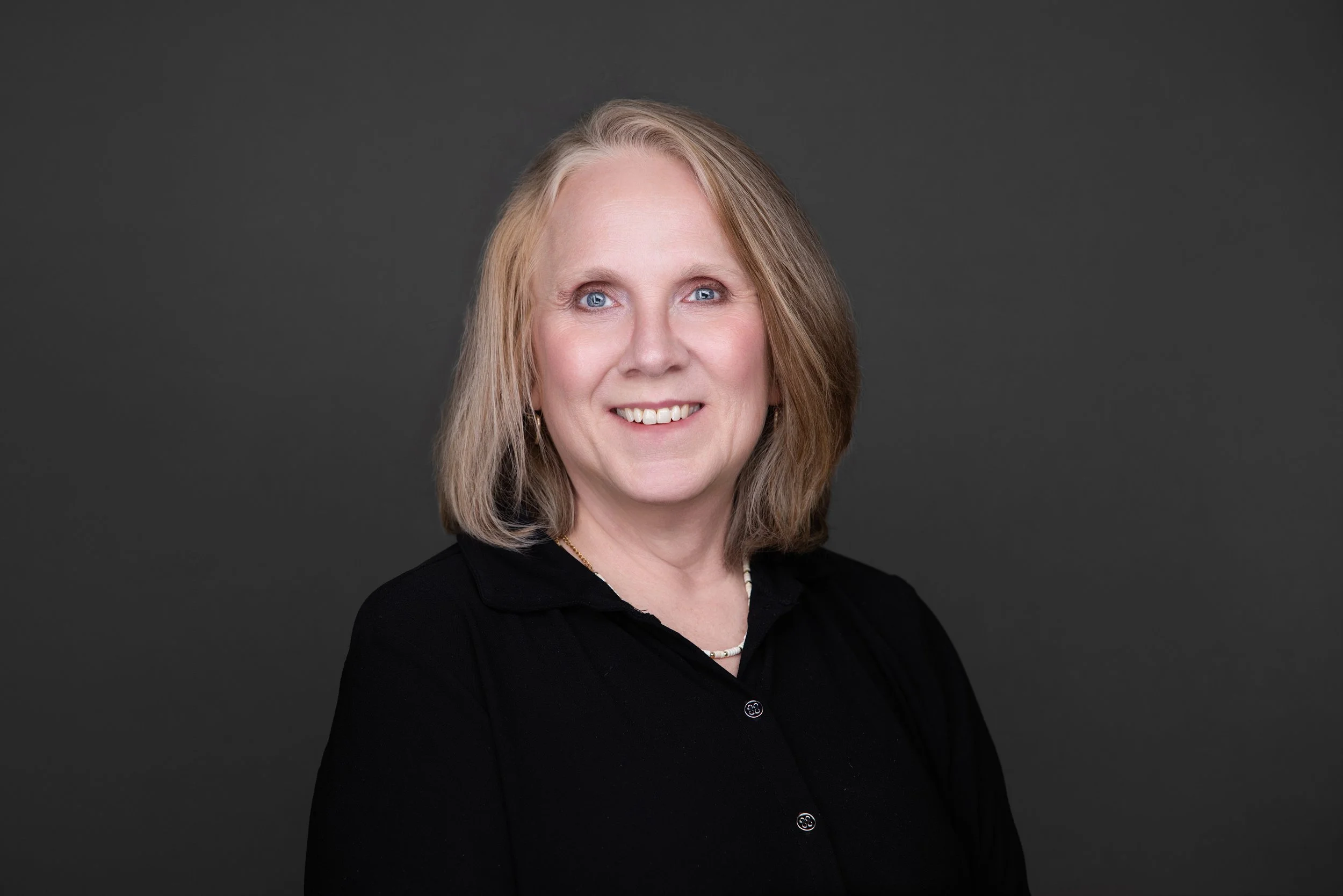 A professional woman with blonde hair, blue eyes, and a friendly smile wearing a black top against a dark gray background.
