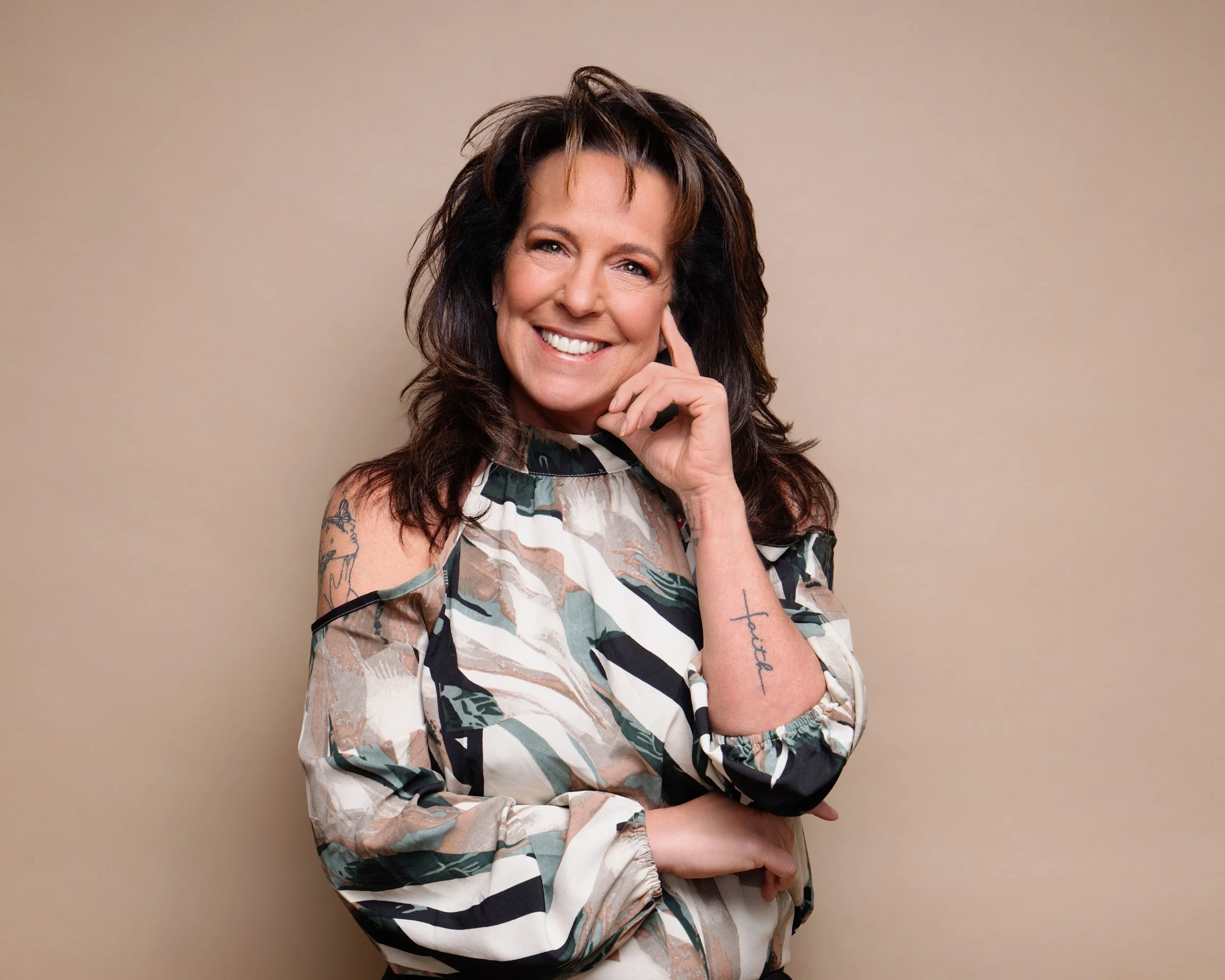 A woman with dark, wavy hair smiling and touching her face, wearing a patterned cold-shoulder blouse with a beige background.