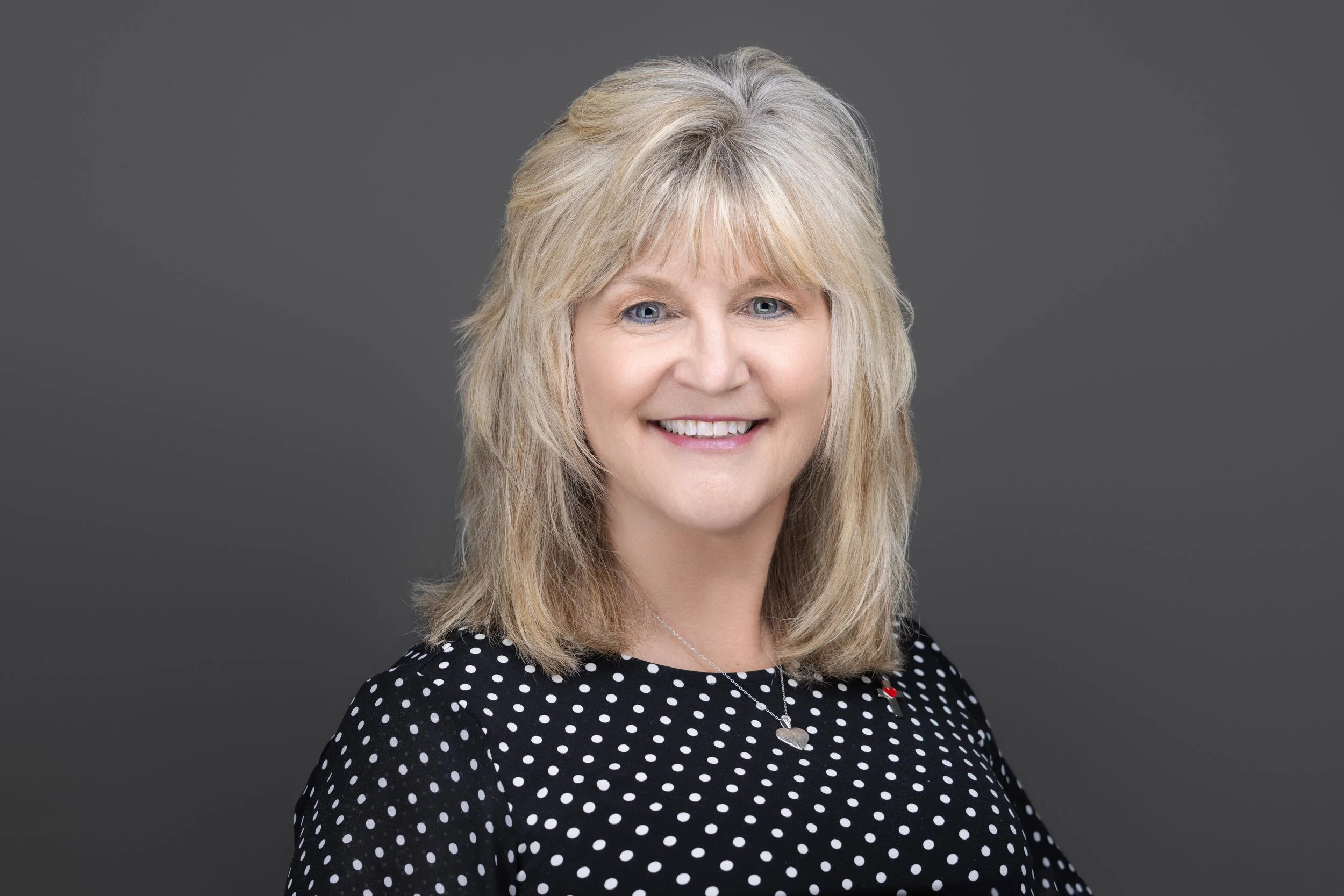 A smiling woman with blonde, shoulder-length hair wearing a black polka dot blouse and a delicate necklace, standing against a gray background.