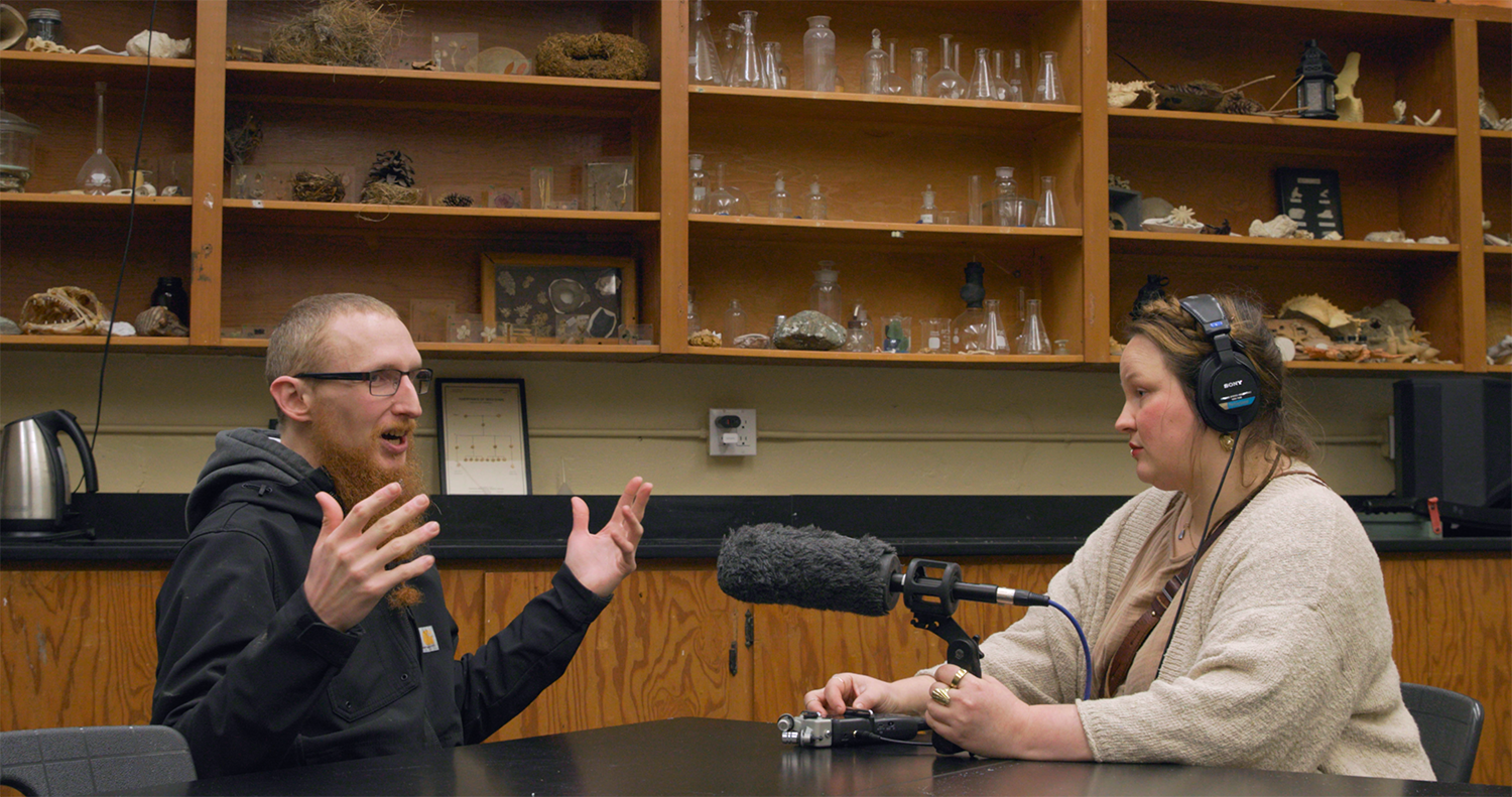 A woman wearing headphones sits at a table, speaking into a microphone held by a person across from her, who is gesturing with his hands.