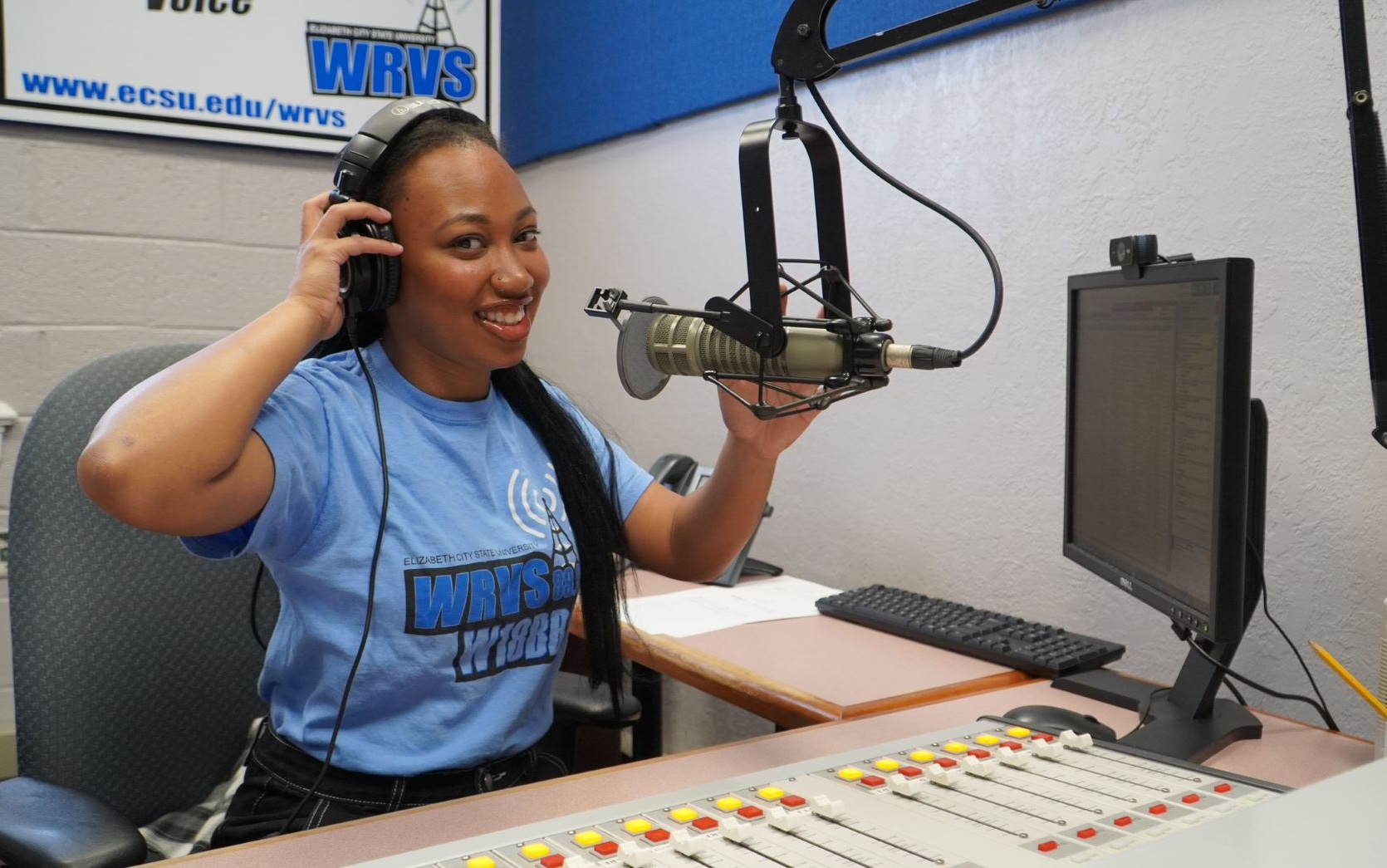 A woman with long black hair wearing a blue WRVS T-shirt, sitting at a radio station in front of a microphone, headphones on, smiling, with a computer monitor, soundboard, and a WRVS banner in the background.