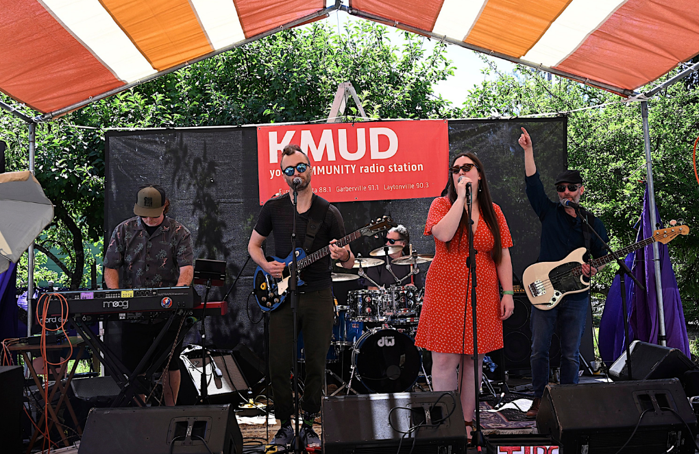 A band of five members performing on an outdoor stage under a striped canopy with trees in the background. The band includes a keyboard player, a guitarist, a drummer, a female vocalist, and a bass guitarist. A large red banner behind them reads 'KMUD' and 'your COMMUNITY radio station'.