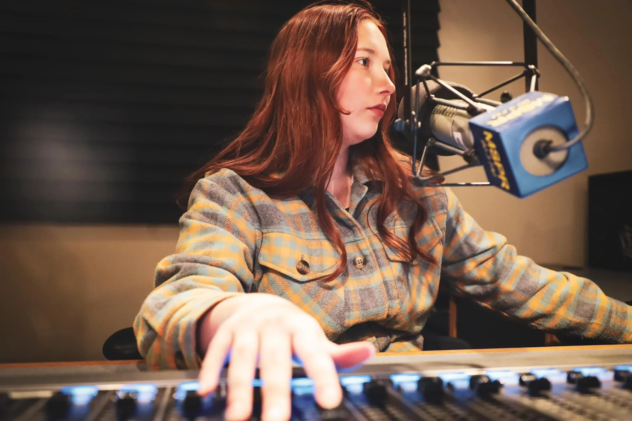 A woman with long red hair wearing a plaid shirt, operating a radio station microphone and soundboard in a recording studio.