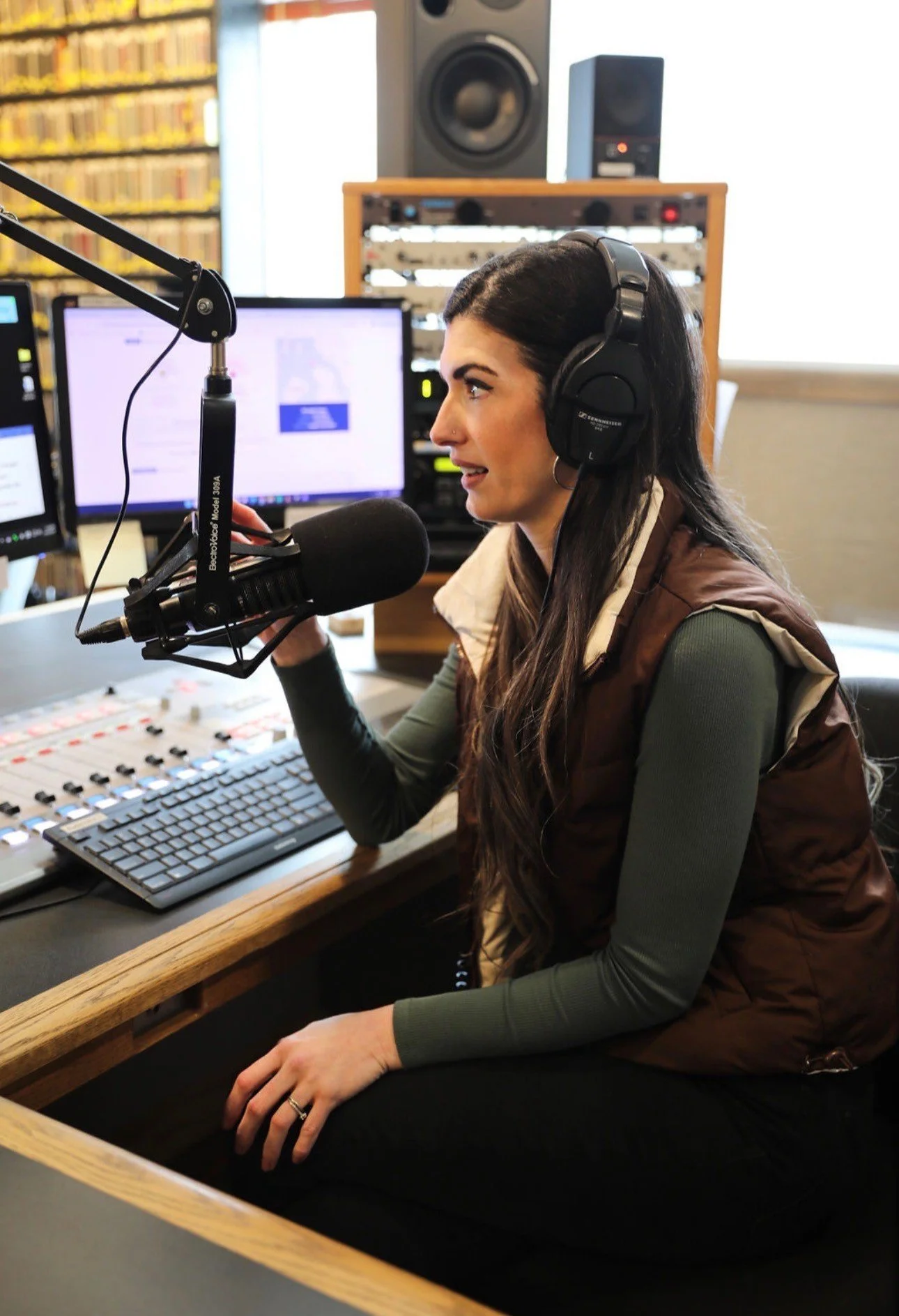 A woman speaks into a professional broadcast microphone in a radio studio, wearing over-ear headphones. Audio equipment and a computer monitor are visible in the background.