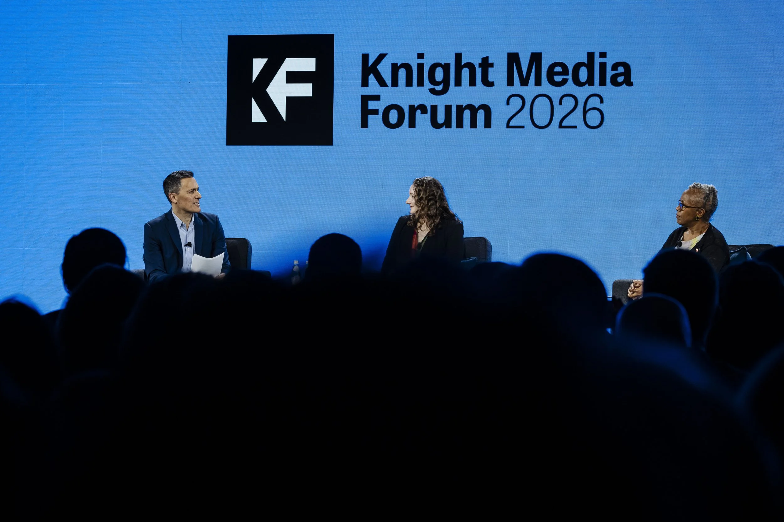 Three people sit in black chairs onstage at the Knight Media Forum. A big blue wall reads "Knight Media Forum 2026," and silhouettes of the audience are in the foreground.