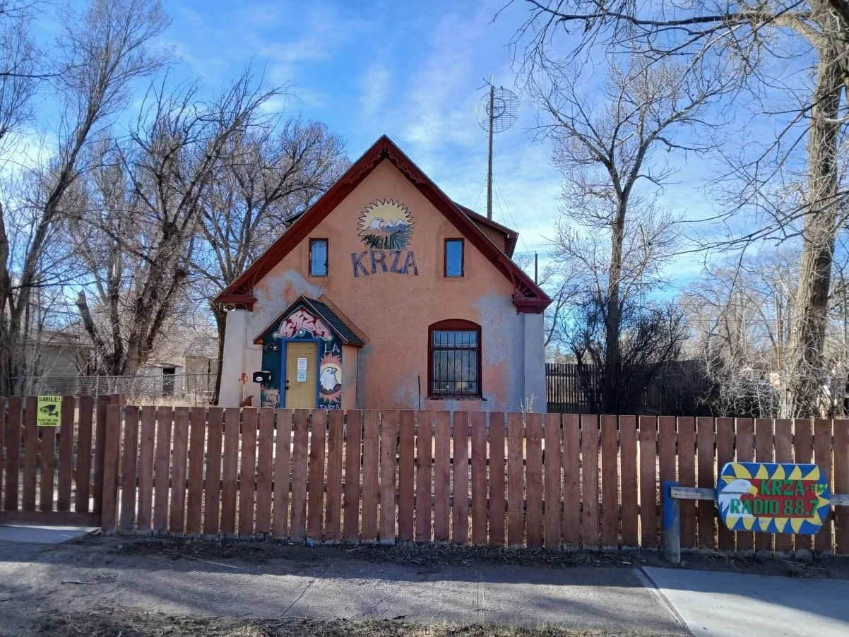 A light orange building is pictured, with a red roof and the logo for radio station KRZA is pictured against a blue sky backdrop and a wood picket fence in the foreground.