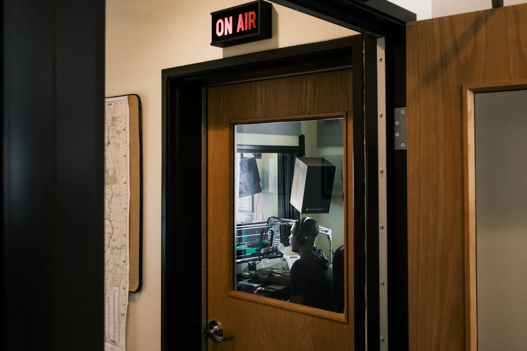 Radio station studio with a person wearing headphones working at a soundboard and computer monitors. An 'ON AIR' sign is lit above the door.